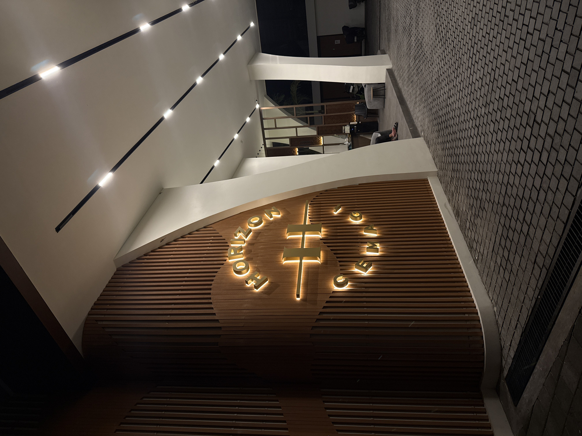 The image presents a modern architectural interior, likely a lobby, atrium, or entrance area, viewed from an elevated perspective, possibly a balcony or upper floor. The setting appears to be at night or evening, as evidenced by the reliance on artificial lighting and dark backgrounds through openings.
The dominant feature is a large, curved wall faced with horizontal wooden slats, which serves as a backdrop for a prominent, glowing golden sign. At the center of this sign is a stylized, illuminated golden cross. Surrounding the cross are glowing golden letters, which spell "HORIZO" on the left side and "GEMMA" on the right side, suggesting the full word "HORIZON" on the left.
The space is illuminated by bright, linear recessed lights embedded in the white ceiling, creating a clean, modern aesthetic. The left side of the frame is dominated by these white architectural elements, including walls and ceilings that curve and intersect.
On the right side of the image, a dark, rectangular paved floor is visible, extending into the background. In the mid-ground, a person is seated on a chair at a table in a lounge or waiting area, their feet visible. The area behind them features modern furniture and a railing with glass partitions, hinting at a multi-level structure or an open-plan design. The overall atmosphere is sophisticated and inviting, with a mix of warm wooden textures and cool, clean lines.