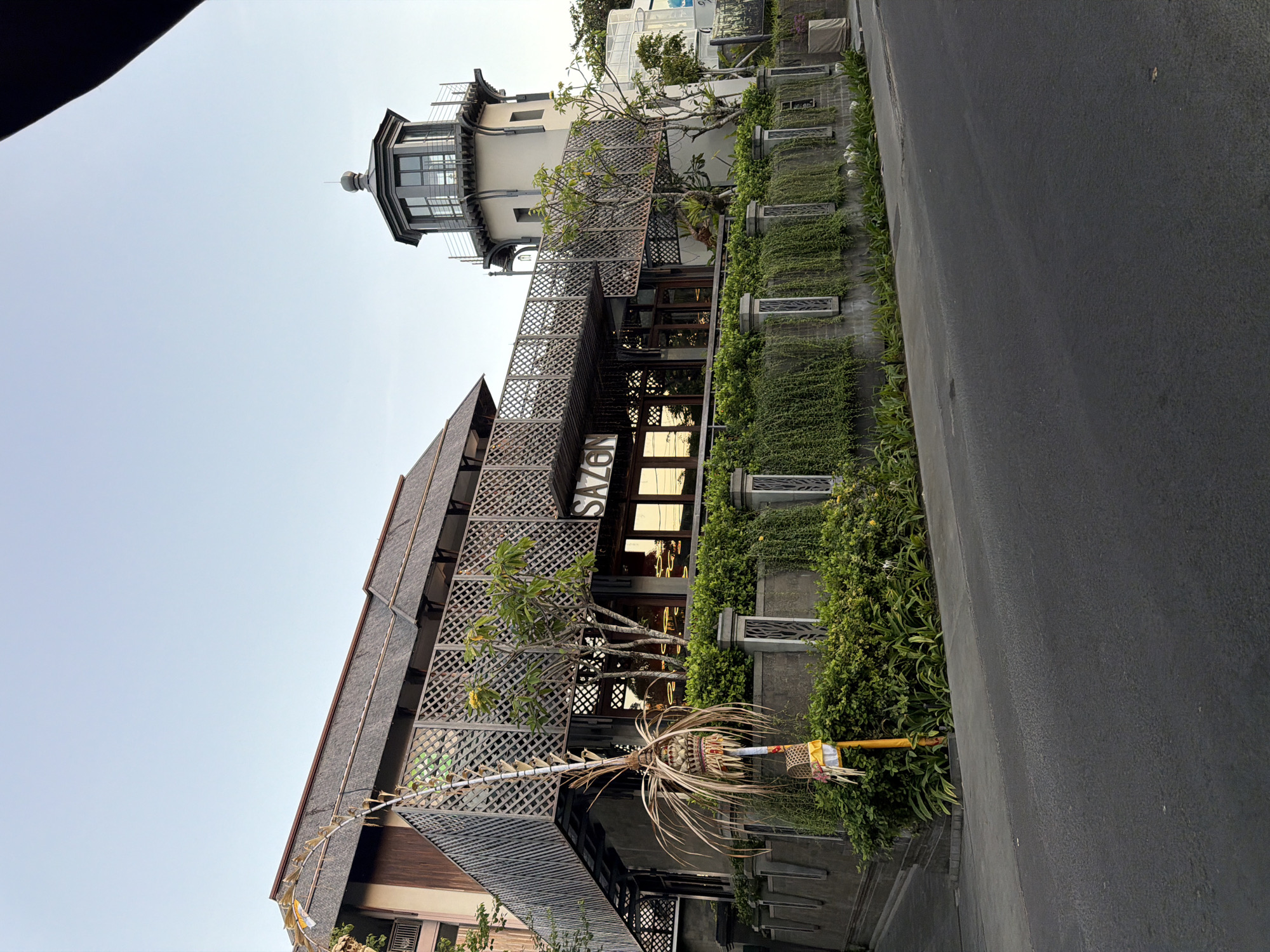 This is a medium-angle, eye-level, outdoor, full shot of the Sazen Restaurant located in North Kuta, Indonesia. The restaurant, with its lighthouse-like tower, is set behind a gray stone wall adorned with lush green hanging plants and decorative pillars. The building features a traditional Balinese thatched roof and intricate wooden lattice work on its balconies. A tall, decorative bamboo pole with offerings stands in front of the building. The sky is clear and a pale blue. The foreground shows a dark asphalt road with a concrete sidewalk. There are no people visible in the image.
