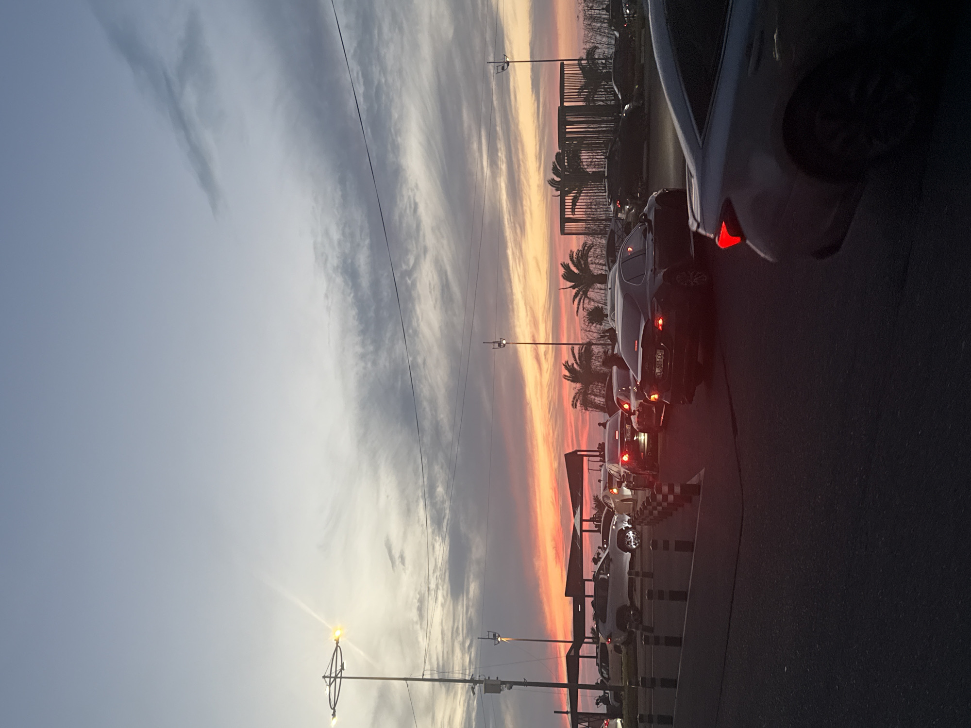 The image shows a sunset over a street in Batumi, Georgia, with a line of cars stopped in traffic. The sky is a mix of gray, white, orange, and red hues. Palm trees and streetlights are visible along the road. The cars are mostly dark-colored, with red taillights illuminated. The road surface is asphalt.