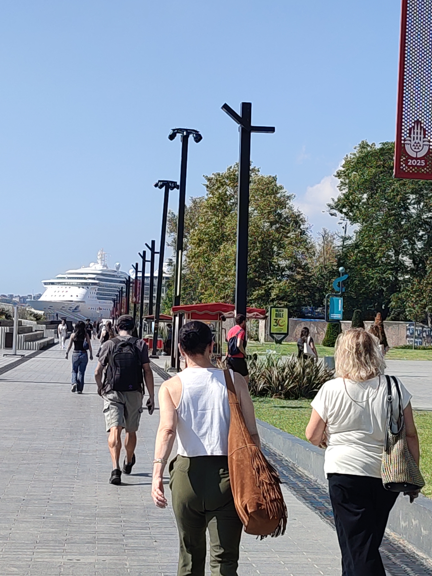 The image depicts a sun-drenched promenade in Beyoğlu, Turkey, likely near the Galataport cruise terminal. A large, white cruise ship is docked in the background, its multiple decks and balconies visible against a clear blue sky. The promenade is paved with grey cobblestones and lined with modern lampposts. Several people are walking along the promenade. In the foreground, two women are walking away from the camera. The woman on the left has blonde, curly hair and is wearing a white t-shirt and black skirt, carrying a woven shoulder bag. The woman on the right has her back to the camera, wearing a white tank top and green pants, carrying a large, fringed brown bag. Further down the promenade, a man with a backpack and a man in a grey t-shirt and khaki shorts are walking away from the camera. Other individuals are visible further in the distance, and there are trees and green spaces to the right of the promenade. Red awnings of small kiosks or cafes are visible on the right side of the image. A banner with the year "2025" and a symbol is visible in the upper right corner.