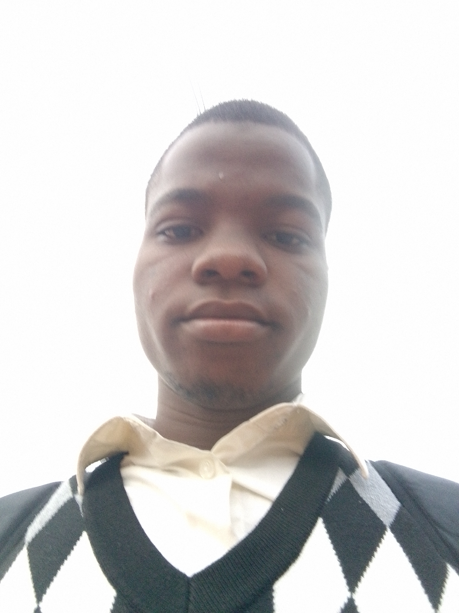 A low-angle, close-up portrait shows a Black man with short, cropped hair. He has a neutral expression. He is wearing a light-colored collared shirt under a black and white argyle sweater. The background is overexposed and white. The location, Osu, Nigeria, cannot be determined from the image alone.