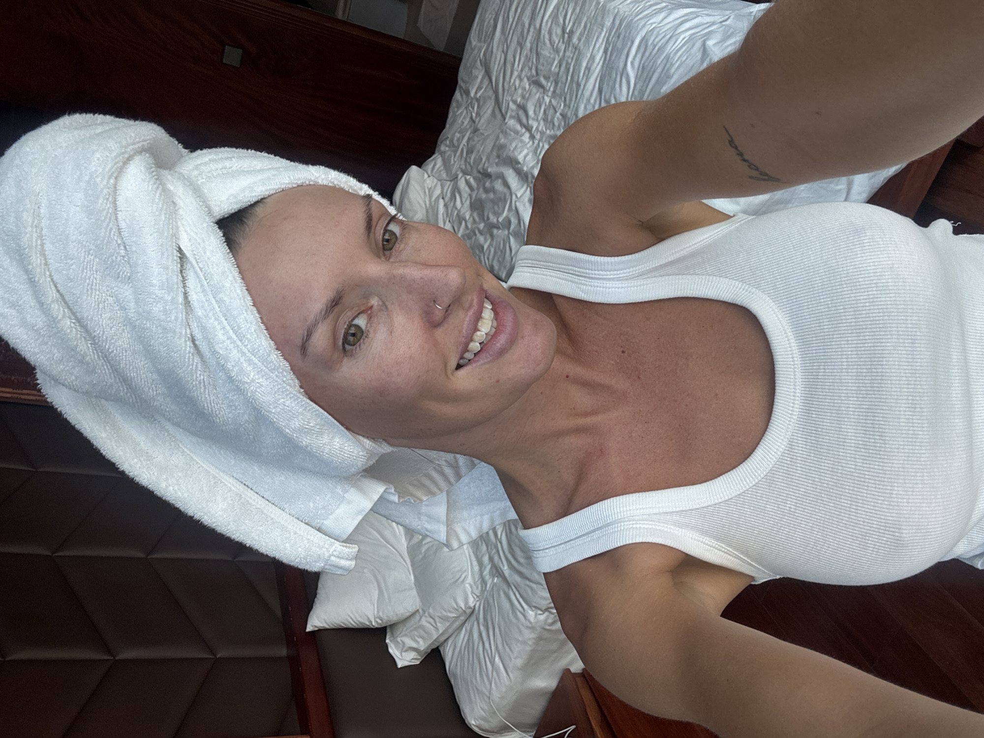 The image shows a woman in a white tank top and a white towel wrapped around her head. She has a nose ring and a tattoo on her arm. She is lying in a bed with white bedding. The headboard of the bed is brown. The image does not contain sufficient information to ascertain its location in Đà Nẵng, Vietnam.