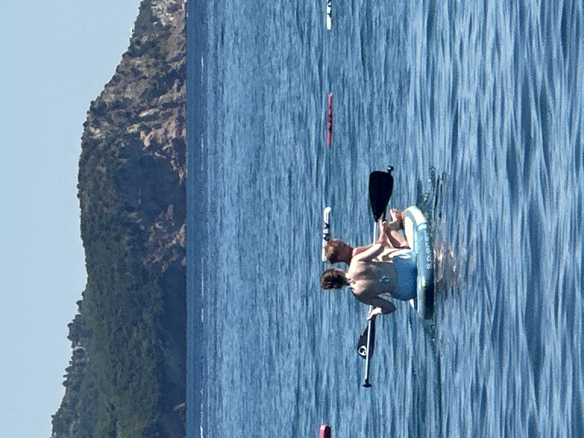 This media file captures two people engaged in stand-up paddleboarding on a body of water, with a lush, green, rocky hillside in the background. The water is a deep blue, with visible ripples and texture, suggesting a breezy day. The sky above is a pale blue.
The two individuals are on a paddleboard, with one person sitting and the other appearing to be kneeling or leaning. They are both shirtless, wearing swim trunks. One of the individuals has a paddle with a black blade resting beside them, and another paddle is partially visible in the water. Another paddleboard, a bright red one, is visible further in the distance, as well as a white one.
The scene is set on a sunny day, with the light suggesting it could be late morning or early afternoon. The rocky, vegetated cliff face on the left side of the image provides a sense of a coastal or lakeside environment. The activity is recreational, indicating a pleasant and active day out on the water. There is no visible text in the image. The overall mood is one of leisure and enjoyment of the outdoors.