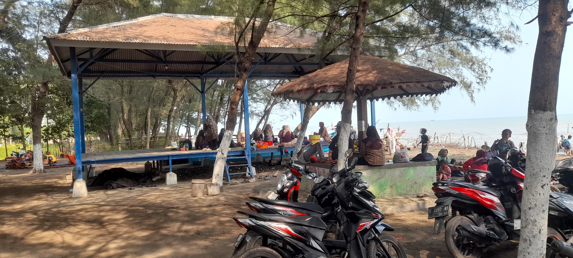 The image depicts a beachside scene in Nyamplung Sari, Indonesia, with two covered structures. The larger structure on the left is an open-sided pavilion with a corrugated metal roof and a blue-painted wooden platform. Several people are seated on the platform, some appearing to be families. To the right, a smaller, circular pavilion with a thatched roof provides shade. In the foreground, several motorcycles are parked, with two prominently displayed. The beach area beyond the pavilions shows the ocean and a light blue sky. Trees are present throughout the scene, providing shade. In the distance, the outlines of small boats can be seen on the water, and there are some scattered wooden structures along the shoreline. Some children are also visible near the water's edge and playing. The ground is sandy, with some dirt and debris scattered around. The overall impression is that of a relaxed, communal gathering spot by the sea.