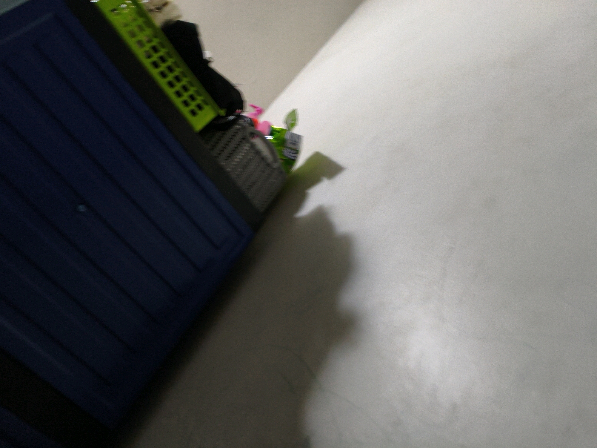 In the city of Karangtalun, Indonesia, an indoor scene is depicted with a blue cabinet on the left and a light-colored wall on the right. The cabinet is partially visible, showing its ribbed surface. On top of the cabinet and slightly to the right, there are several items including a green plastic basket, a dark object, and some colorful bottles or containers, one of which is pink. A shadow, likely cast by an object or person out of frame, is visible on the wall and floor to the right of the cabinet. The lighting suggests an indoor environment, possibly a room in a residence or storage area.
