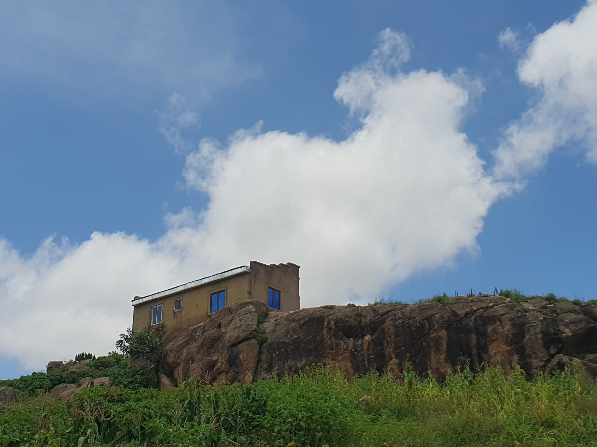 An image taken from below shows a simple, light brown building with a metallic roof situated atop a rocky outcrop in Gura Top, Nigeria. The building has at least three windows, two with blue frames and one with a white frame, and appears to have decorative crenellations along its top edge. The rocky outcrop is covered with sparse vegetation, and the foreground is dominated by lush green plants, possibly crops like corn. The sky above is a bright blue with scattered white clouds. The overall impression is of a remote, elevated dwelling in a natural, possibly rural, setting.