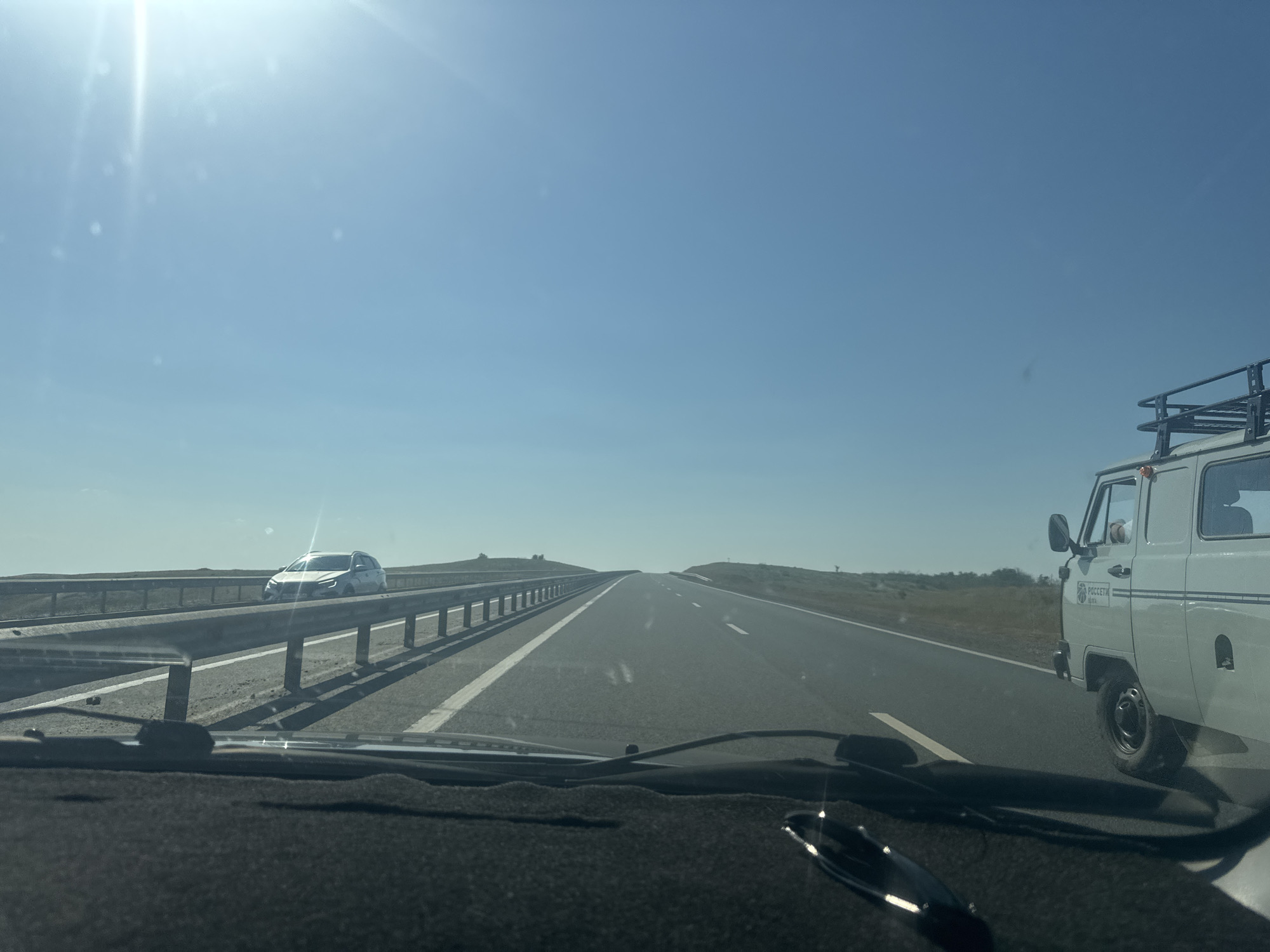 A clear, sunny day is depicted from the perspective of someone inside a vehicle traveling on a highway. The road ahead is paved and divided into lanes by white lines, curving slightly to the right. A metal guardrail runs along the left side of the road, separating it from a grassy embankment. In the distance on the left, a white car is visible on the other side of the guardrail. On the right side of the highway, a light-colored van with a roof rack is traveling in the same direction. The van has some text and a logo on its side. The sky is a bright, cloudless blue, with the sun creating a lens flare in the upper left quadrant of the image. The foreground shows the interior of the vehicle, including the dashboard and windshield. The surrounding landscape appears to be open, with rolling hills covered in dry vegetation. The scene suggests travel through a rural or semi-rural area.