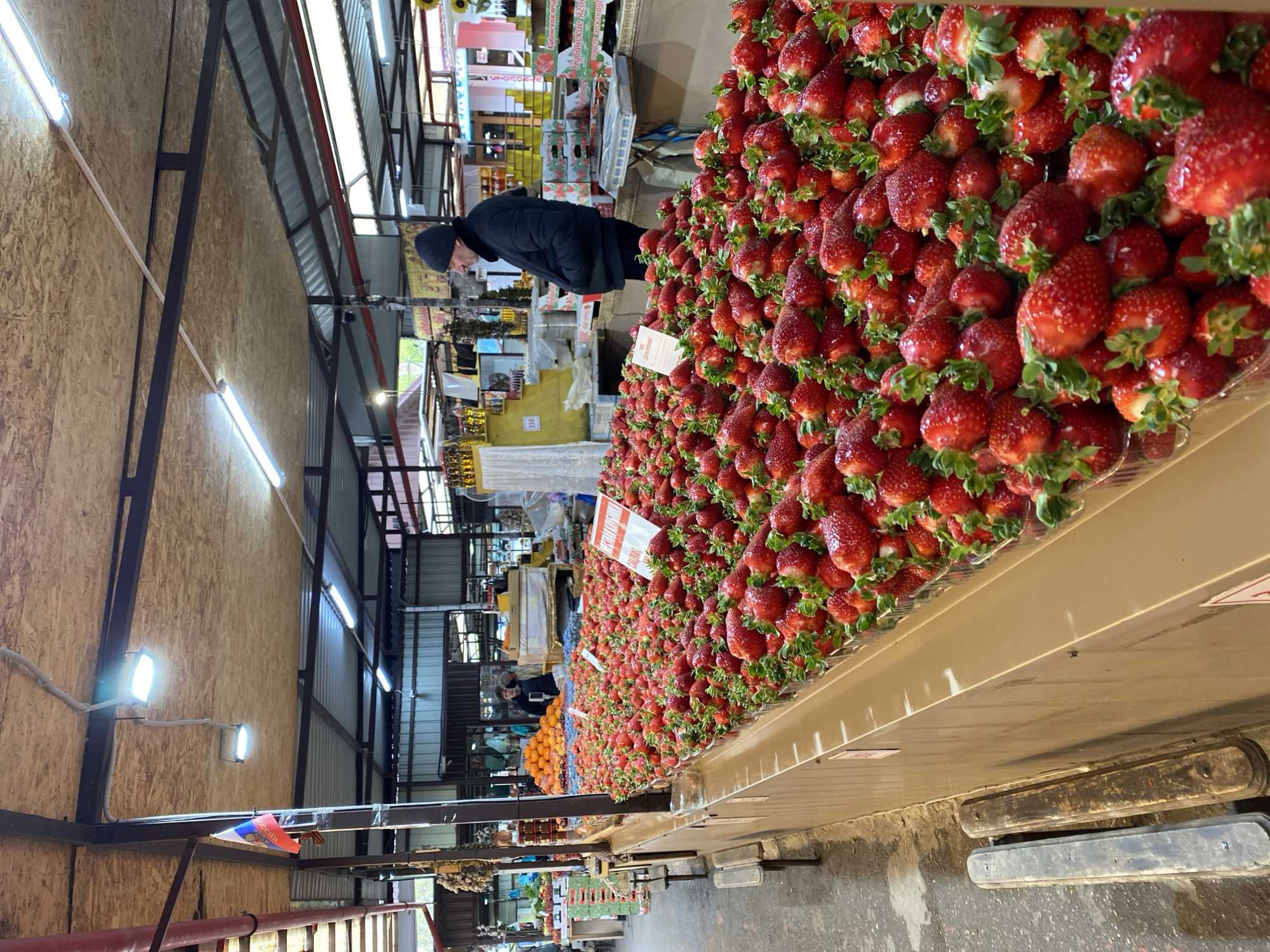A vendor at a market in Gelendzhik, Russia, stands behind a table piled high with strawberries. The man is wearing a black coat and hat and is looking at something behind the table. The strawberries are arranged in plastic containers. Other produce is visible, including oranges. The market appears to be indoors. The walls are paneled and the ceiling has fluorescent lights. A Russian flag is visible in the background.