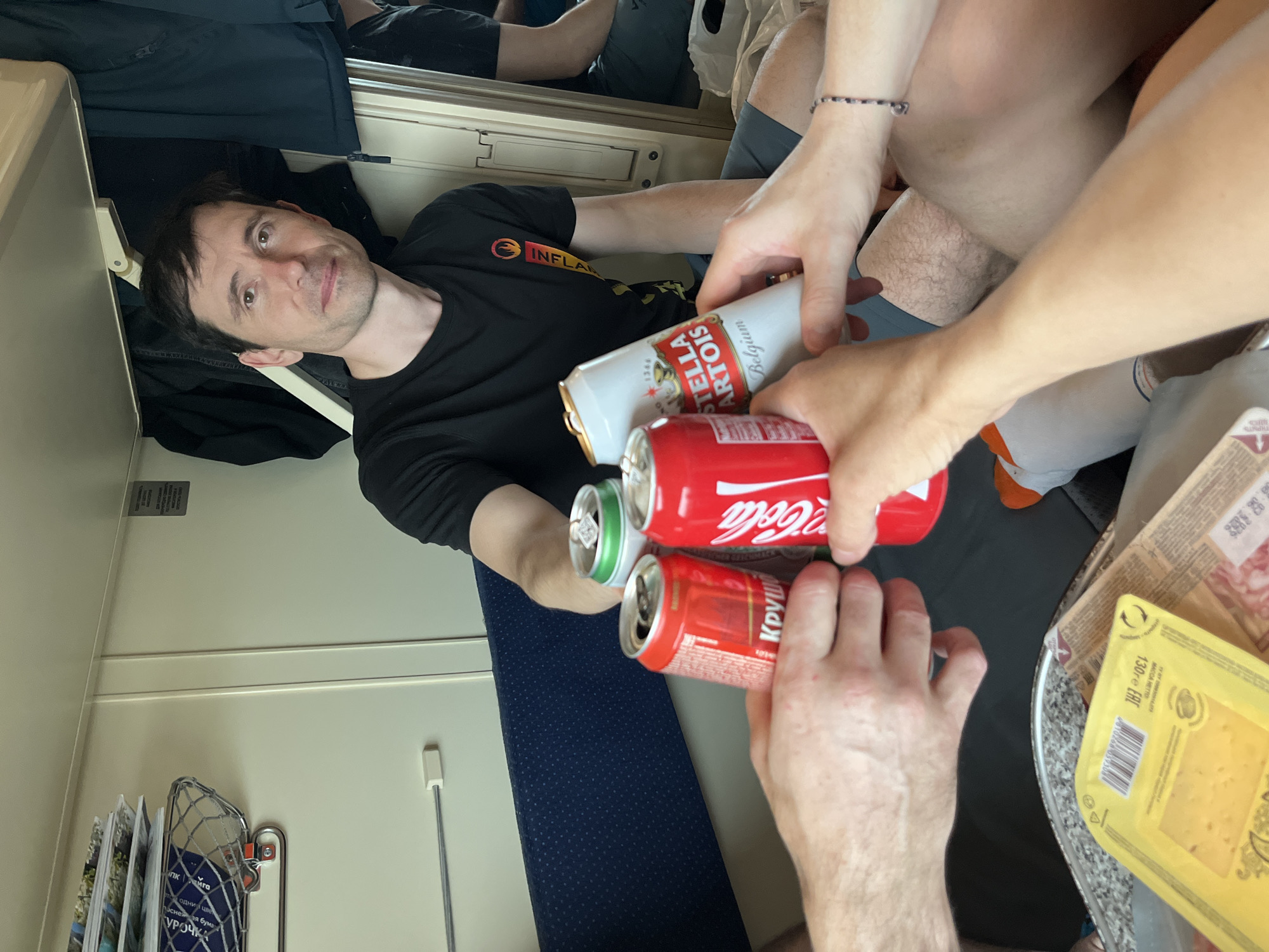 A medium shot shows a toast in a train compartment. A man in a black T-shirt, with "INFLA" visible on it, sits and looks toward the camera while partially holding up a can of Stella Artois. Two other pairs of hands hold cans of Coca-Cola and another can of an unidentifiable red-labeled beverage, creating a toast-like composition. Nearby is a partially visible tray with cheese and meat products. Train details include a magazine rack, and blue seat upholstery. Information on food products in the image is written in Russian. Without further context, the location of the city of Izhevsk cannot be verified.