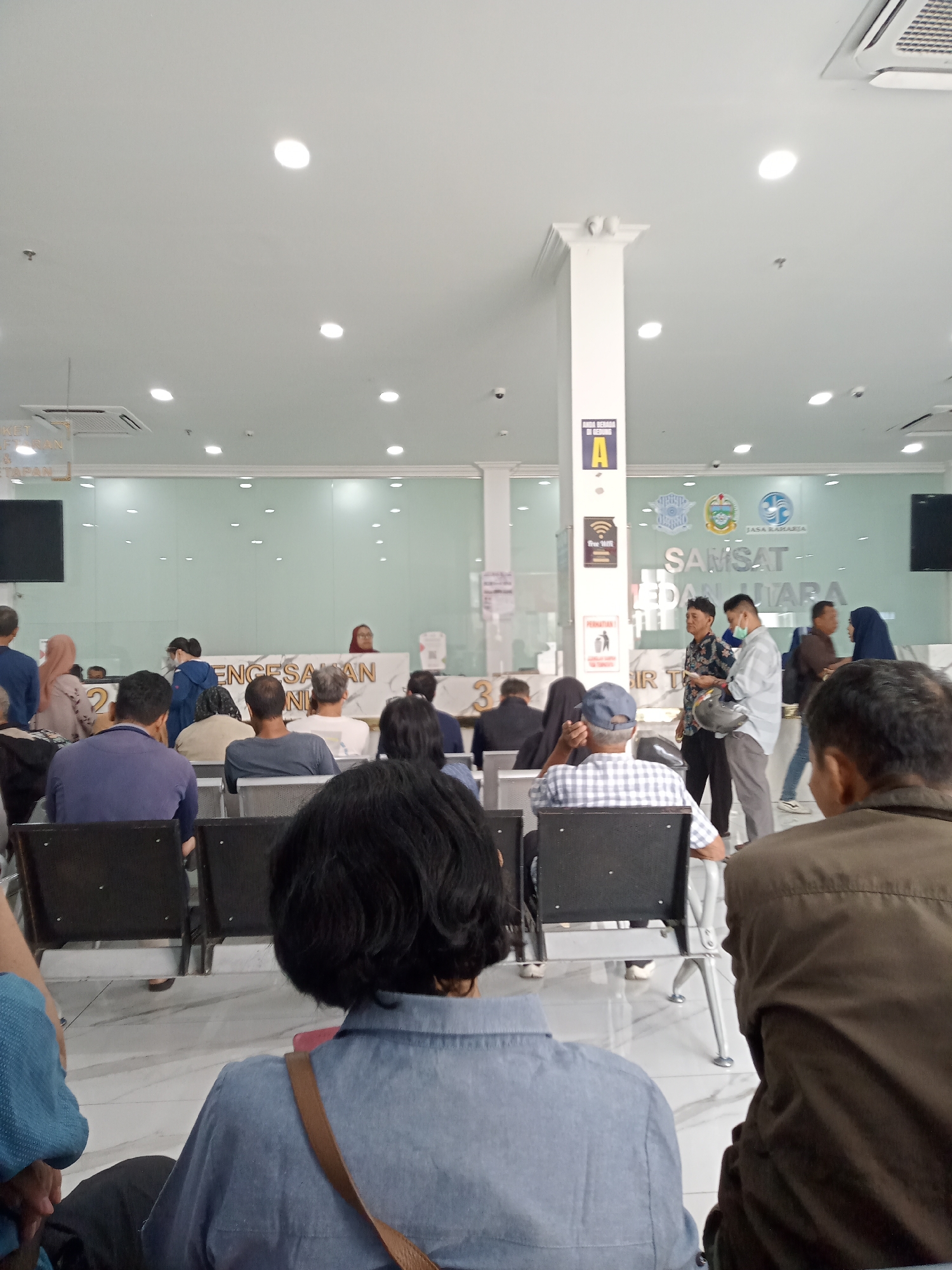The image shows the interior of a government office in Medan, Indonesia, likely the Samsat (Sistem Administrasi Manunggal Satu Atap) office, which handles vehicle registration and taxes. The scene depicts a waiting area with several people seated on benches and standing. A counter with employees is visible in the background. The environment appears to be a typical public service office with fluorescent lighting and a clean, modern aesthetic. The overall impression is one of a busy administrative center.