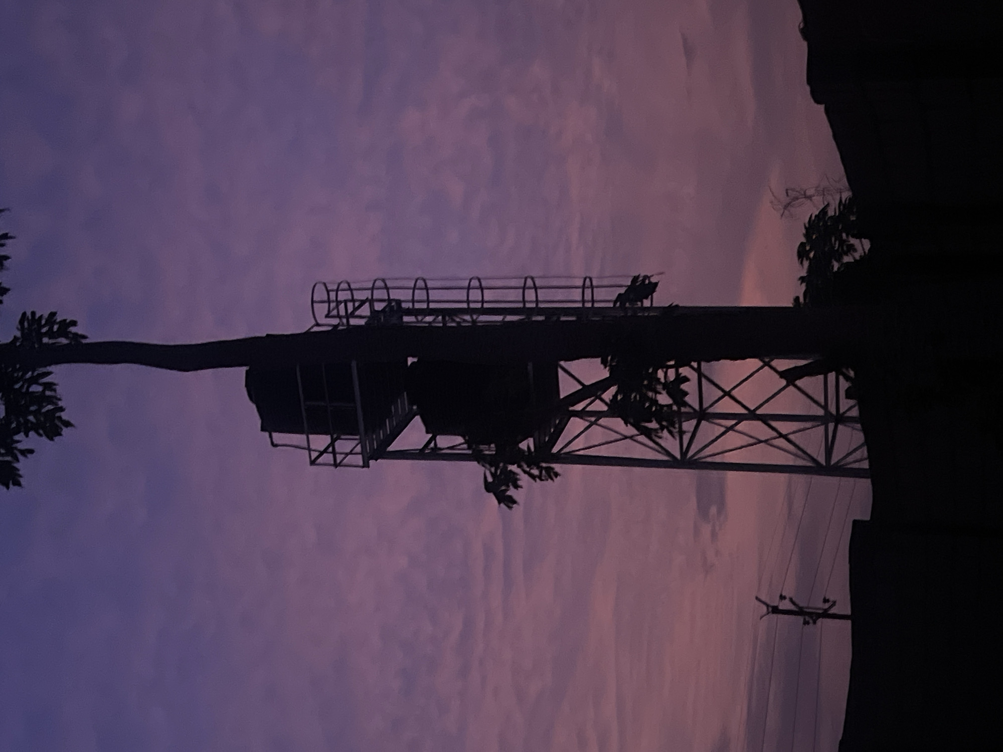 The image depicts a silhouette of a structure, possibly a water tower or telecommunications tower, against a twilight sky with pink and purple hues. The structure is made of metal with a ladder and platform. Trees and power lines are visible in the foreground and on the right side of the structure. The location is Gura Top, Nigeria.