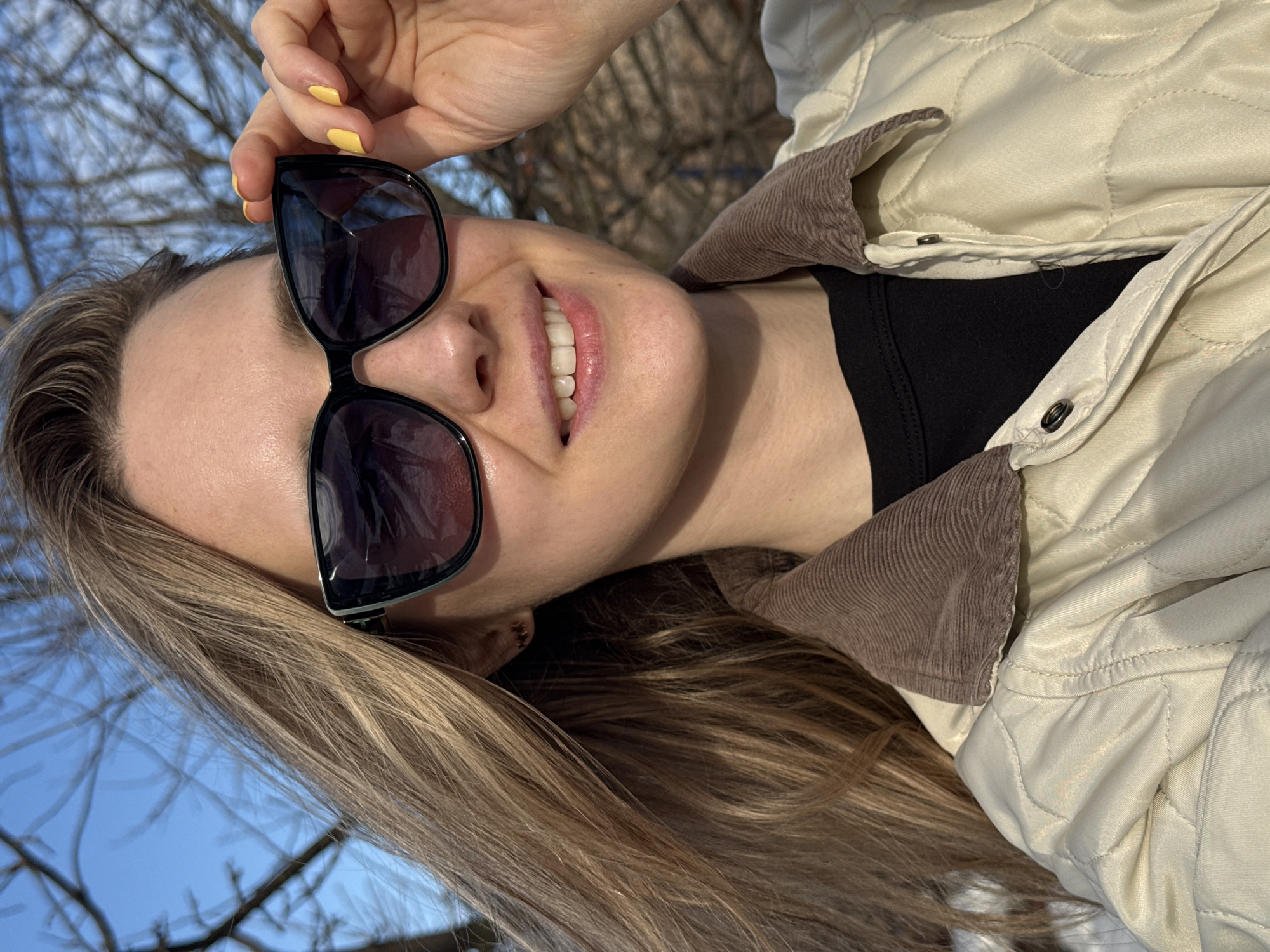 A young woman with blonde hair is pictured outdoors, holding a pair of sunglasses. Her nails are painted yellow. She is wearing a black top and a quilted, off-white jacket with a brown corduroy collar. The background shows a bare tree and a blue sky, suggesting a sunny day in late fall or early spring. The location is Chaikovsky, Russia.