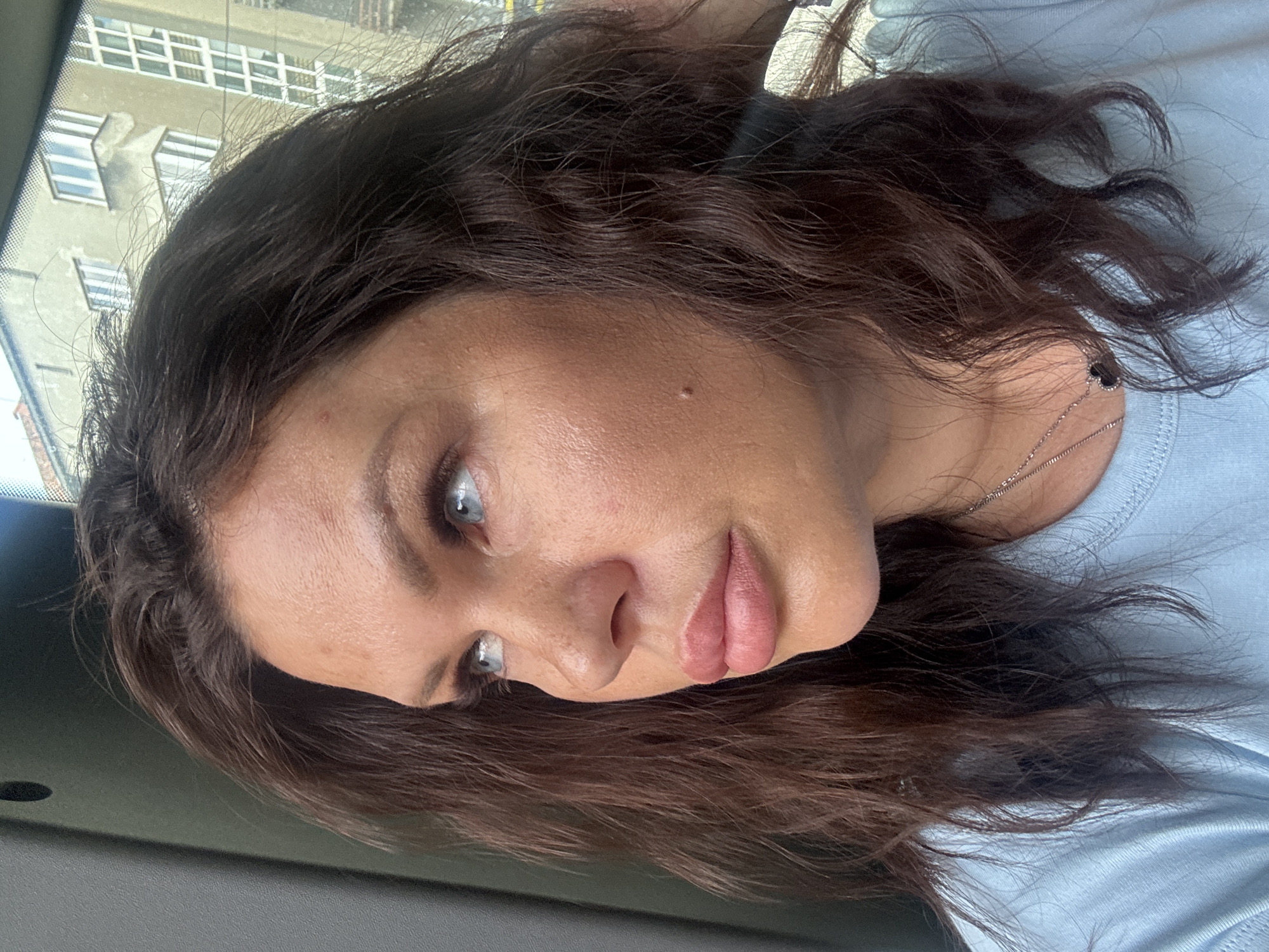 The image shows a close-up portrait of a woman with medium-length, wavy brown hair, light blue eyes, and fair skin. She is wearing a light blue t-shirt and a silver necklace with a black heart pendant. The woman is looking to her left, and her face is illuminated by natural light. In the background, partially visible through a window, is a building with multiple windows, suggesting an urban environment. The scene is captured in a candid manner, with the woman's attention directed away from the camera. Based on the presence of specific architectural styles visible in the background, which are characteristic of post-war modernist buildings common in Katowice, Poland, and the general atmosphere of the scene, it is plausible that this image was taken in the city of Katowice, Poland.