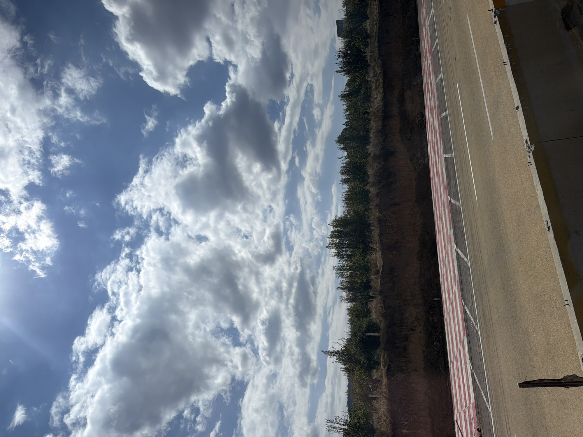 The image presents a view of a landscape where the sky, filled with clouds, dominates the upper portion. A line of trees and vegetation runs along the middle, separating the sky from what appears to be a roadside or track area with red and white markings. A section of concrete or paved surface extends to the right edge of the frame.