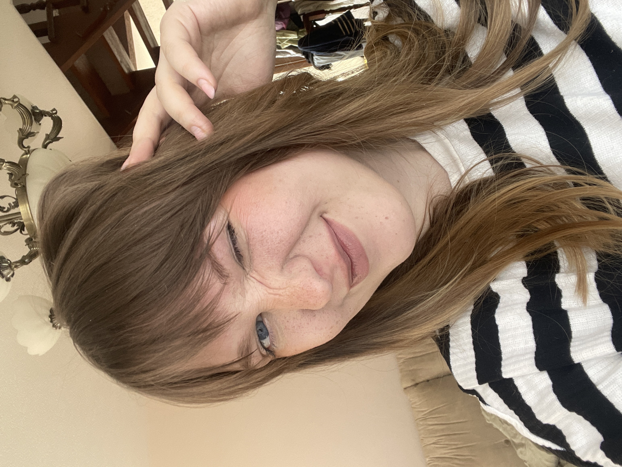 A fair-skinned female with freckles and light brown hair is depicted in a close-up, angled selfie. She is wearing a black and white striped shirt. Her hand is near her head. The background includes a couch and a chandelier. There is no location information available in the image.
