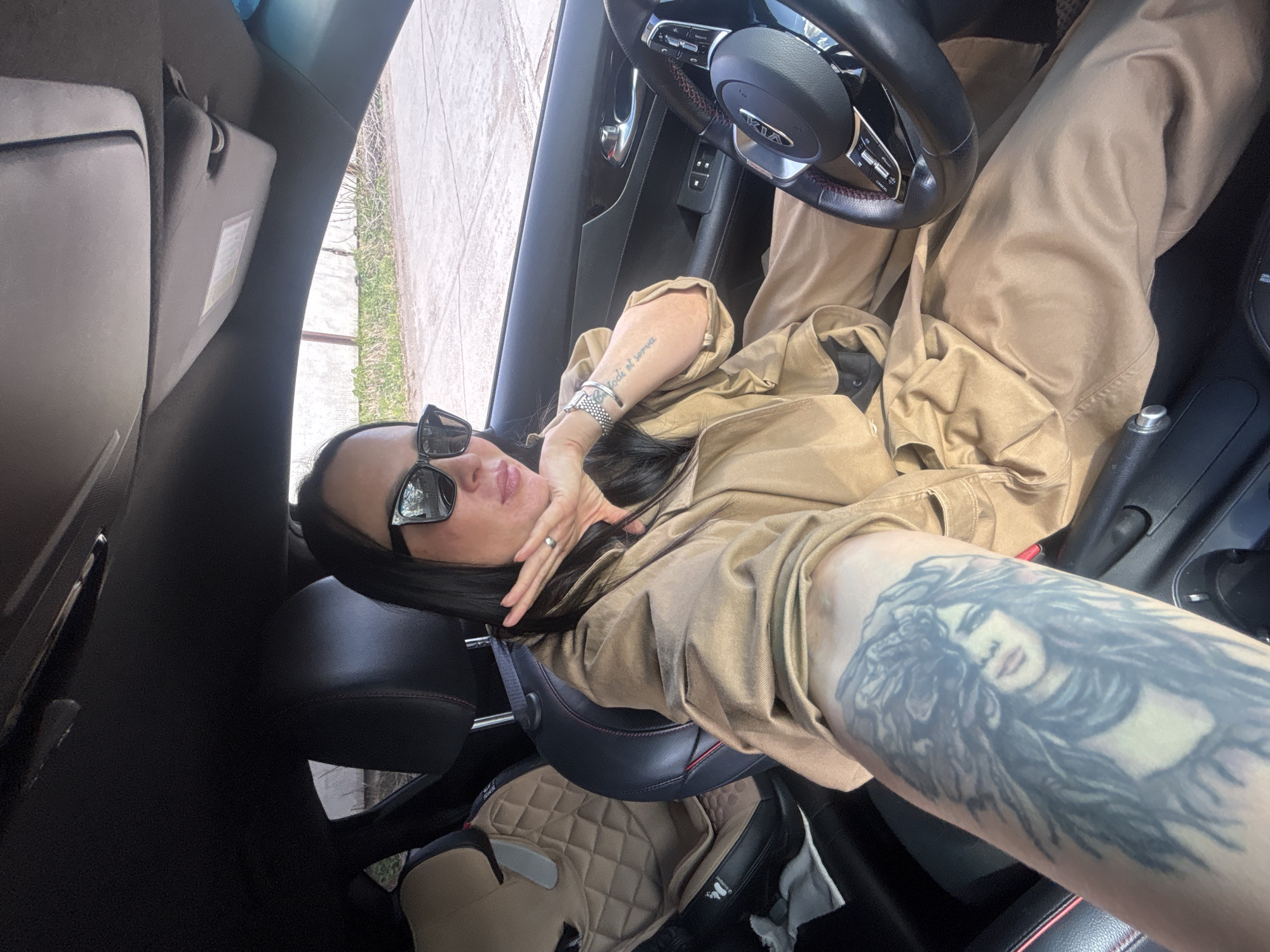 A woman is reclined in the driver's seat of a Kia car. She is wearing a tan jumpsuit and sunglasses. Her left arm, heavily tattooed with a portrait of a woman, is extended towards the camera. She has a watch on her right wrist and is wearing a ring. The steering wheel is visible, and a child's car seat is in the back. The location is an outdoor area in the city of Chaikovsky, Russia, as deduced from the steering wheel.