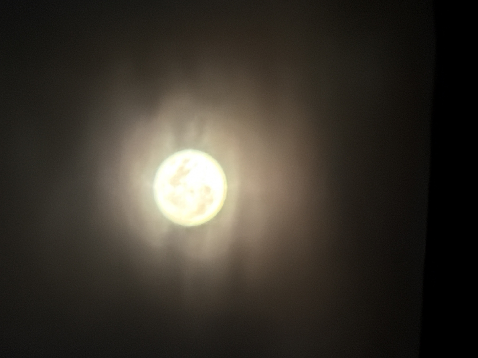 A full moon is visible in a dark sky, surrounded by a soft halo of light. The moon is the brightest object in the image, appearing as a glowing orb with subtle texture. Wispy clouds or atmospheric haze diffuse the moonlight, creating a luminous effect. The lower portion of the image is obscured by a dark, indistinct silhouette, likely representing the horizon or ground. The overall scene is dark and atmospheric, with the moon serving as the sole focal point. No people or specific landmarks are identifiable. The image appears to be a nighttime photograph taken in Kamianka-Buzka, Ukraine.