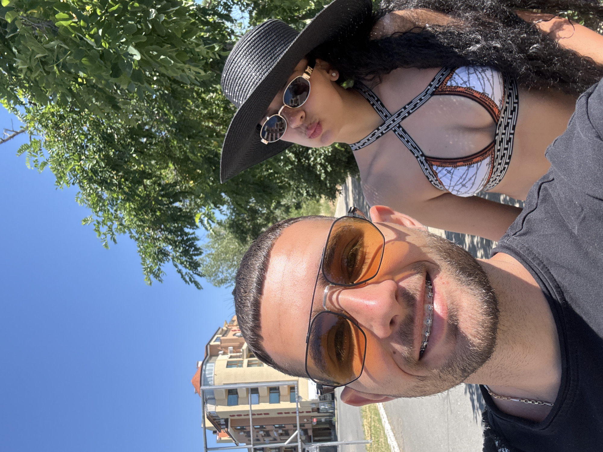 A man and a woman are taking a selfie outdoors on a sunny day. The man, in the foreground and on the left, is wearing a black t-shirt and a silver chain necklace. He has short, dark hair, stubble, and is wearing aviator-style sunglasses with orange lenses and braces on his teeth. The woman is behind him, to the right, and is wearing a black wide-brimmed hat, sunglasses, and a patterned bikini top. She has long, dark, curly hair. In the background, a multi-story building with an orange roof is visible, as well as lush green trees and a clear blue sky. The scene appears to be on a street or promenade in Nesebar, Bulgaria.