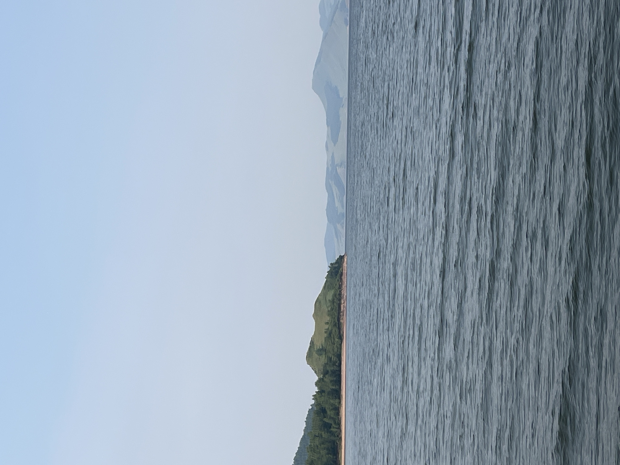 The image shows a wide expanse of calm, dark blue water in the foreground. In the middle ground, a green, rounded hill with a sparsely vegetated summit juts out into the water from the left. The hill is bordered by a sandy shore and a dense forest of green trees on its left flank. Further in the distance, a series of hazy, blue-green mountains form the horizon. The sky above is a clear, pale blue, with no clouds visible. The scene appears to be a natural landscape, possibly a lake or a wide river, near Novoselovo, Russia, given the context provided. No people or man-made objects are visible.