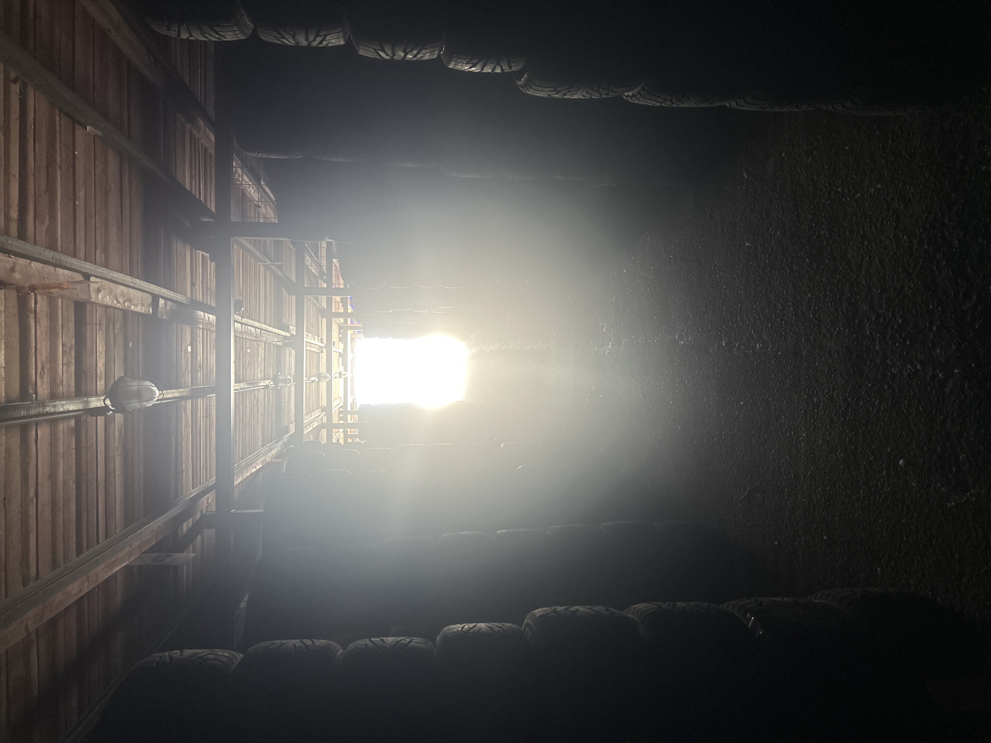 The image depicts the interior of a dimly lit warehouse or storage facility, possibly an industrial building. Tall stacks of dark-colored tires are arranged along the left and right sides of a central aisle, creating a tunnel-like effect. The aisle is covered with a dark, granular material, likely gravel or dirt. The ceiling is constructed of wooden planks supported by metal beams, with industrial light fixtures visible. At the far end of the aisle, a bright light source, possibly a window or an open doorway, creates a strong flare and illuminates the space with a hazy, diffused light, casting long shadows. There are no people or animals visible in the scene. The overall atmosphere is one of storage and disuse, with the bright light at the end suggesting an exit or an area with more activity. The image does not provide any specific clues to pinpoint its location within the city of Saint Petersburg, Russia.