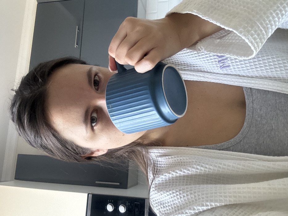 A frontal close-up image depicts a person, appearing female, in a kitchen in Селекционер-2 Снт, Russia. The individual, with dark brown hair, is visible from the chest up, looking directly at the camera. They are holding a teal blue, vertically ribbed ceramic mug with their right hand to their mouth, partially obscuring the lower half of their face. The person is wearing a white waffle-knit robe over a gray tank top; a light purple letter "M" is embroidered on the robe's left lapel area. The background features light-colored walls, dark grey overhead kitchen cabinets, a black appliance with two circular controls integrated into a cabinet on the left, and a white subway tile backsplash on the right.