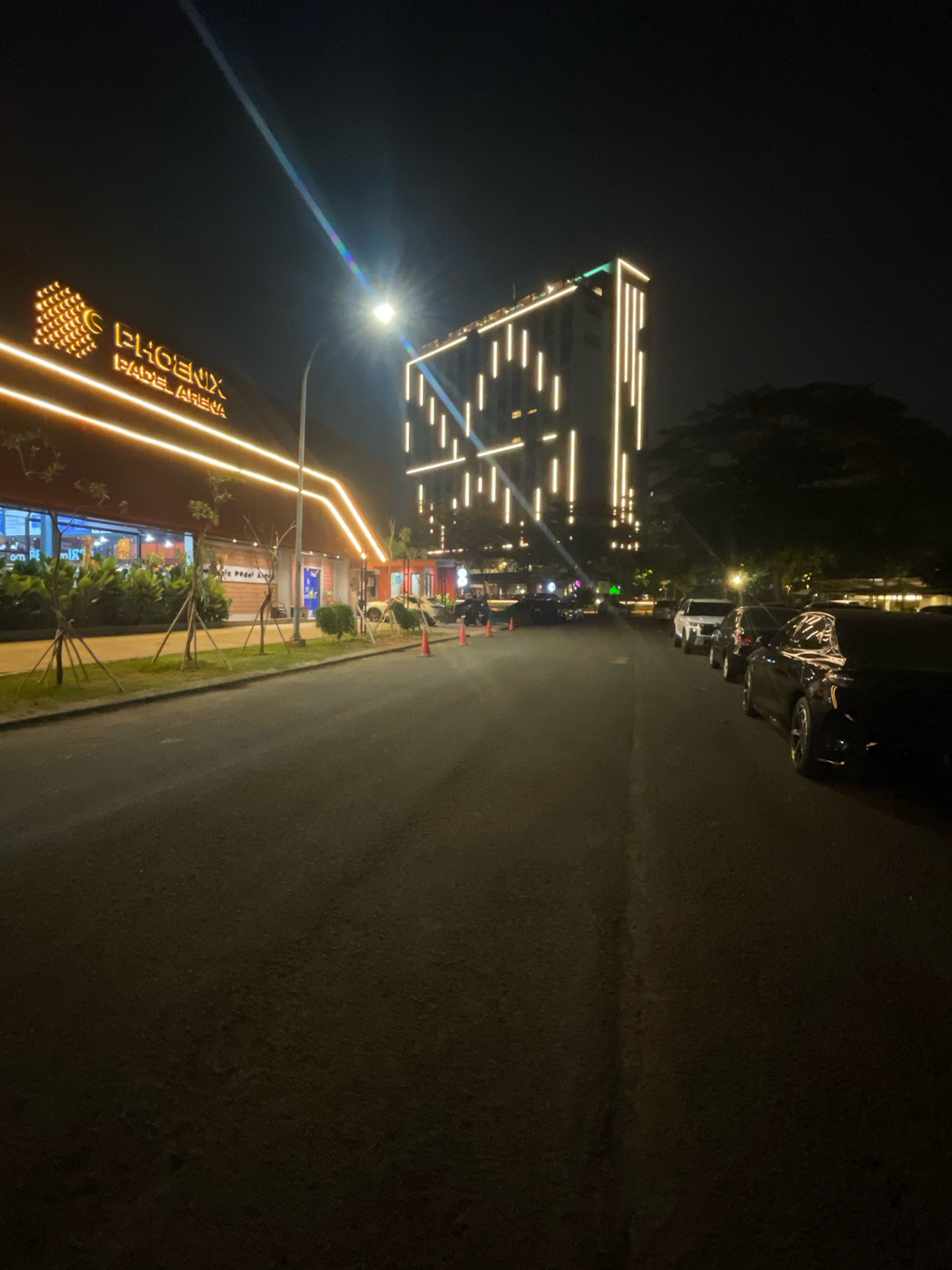 A night-time street view in South Tangerang, Indonesia shows a modern building with illuminated vertical lines, a sign reading "PHOENIX PADEL ARENA" with orange neon lighting, and several parked cars. The street is paved, and the surrounding area is dimly lit, with trees visible in the background.
