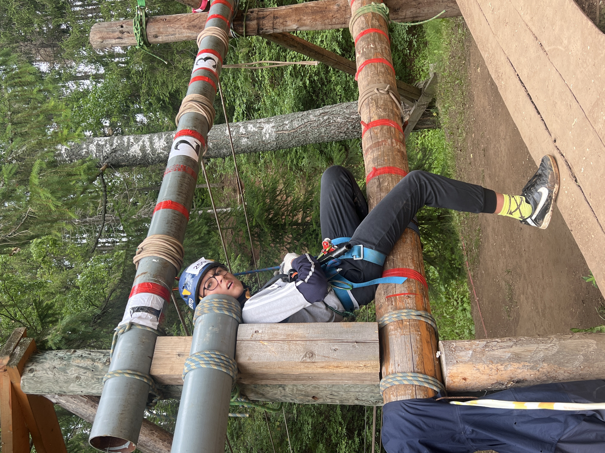A person wearing a helmet and safety harness is suspended between two vertical poles that are part of an outdoor obstacle course. The poles are made of wood and metal pipes, wrapped with ropes and marked with red tape and numbers like "3" and "2". The person is lying on their back, with their legs extended and one foot resting on a wooden beam. They are wearing dark pants, a grey hooded jacket, and black sneakers with bright yellow socks. Their expression is one of mild surprise or amusement.
The background shows a dense forest with tall trees, indicative of a natural setting. The ground appears to be dirt. The overall atmosphere suggests a recreational activity or a challenge, possibly a ropes course or adventure park. The lighting suggests it is daytime, and the foliage indicates a temperate climate. The text "259" is visible on the person's helmet.