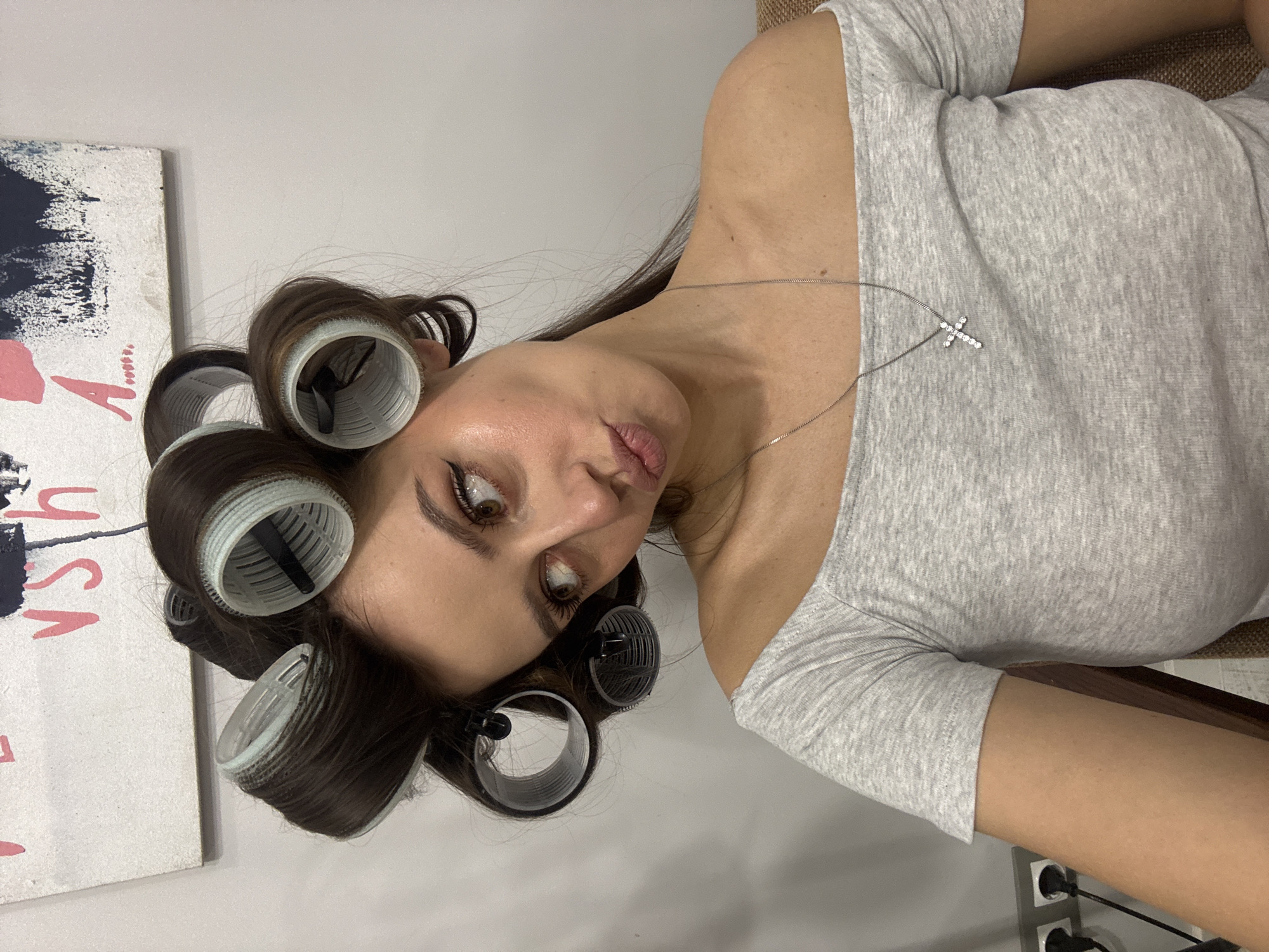 A female individual is depicted in a medium-close-up indoor setting. Her dark brown hair is set in multiple light grey/translucent plastic hair rollers, secured with black clips, visible across her scalp. Her head is tilted to the left, and her gaze is directed towards the upper right of the frame. Her lips are pursed in a distinct expression. She wears visible eye makeup, including winged eyeliner, and has defined eyebrows. Her attire consists of a light grey, off-the-shoulder top or dress. A thin silver-colored necklace with a small, cross-shaped pendant adorned with clear stones rests on her chest. The background features a plain light grey wall. A white canvas or board with abstract pink/red lettering (including a "V" and partial "S" or "H") and dark blue/black brushstrokes is partially visible on the wall behind her upper head. To the lower left, a segment of a brown wooden furniture piece is present, beneath which two white electrical wall sockets are mounted, containing black plugs and associated wiring. The specific location within Krasnoyarsk, Russia, cannot be determined from the image.