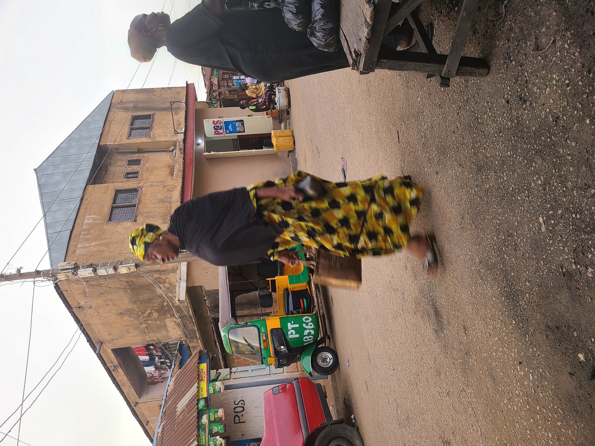 A street in Jos, Nigeria. A woman wearing a black shirt and a yellow and black patterned skirt walks on the street. A green and yellow auto rickshaw with the license plate "PT-18360" is parked on the side of the road. There's a two-story building with a grey roof and tan walls on the left. Various businesses and shops are visible along the street, with signs indicating POS (Point of Sale) services. Overhead electrical wires are visible against a light sky. Another person stands to the upper right in a black robe.