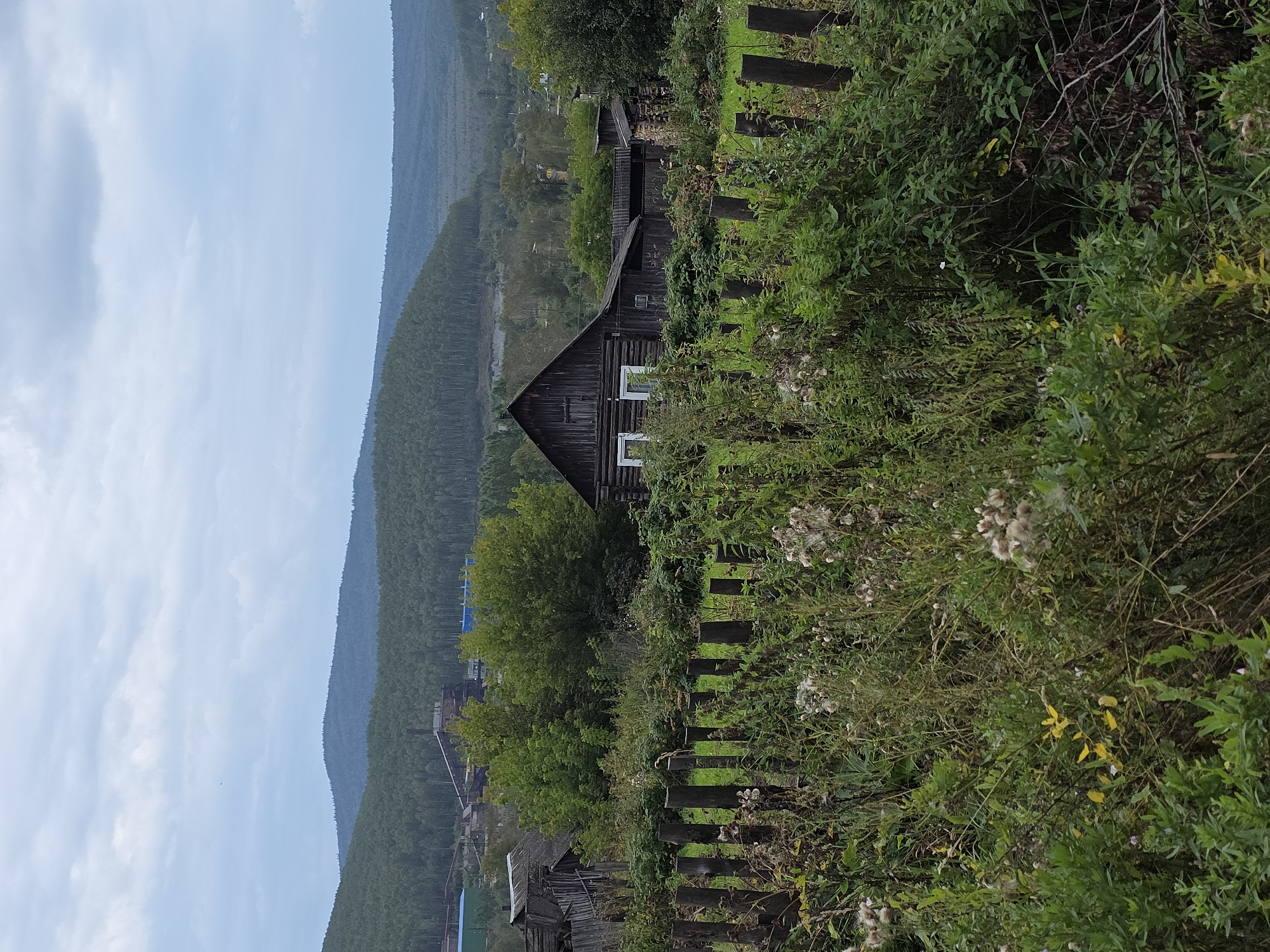 The image depicts a rural scene in the city of Magnitka, Russia, characterized by a wooden house and surrounding overgrown vegetation. A rustic wooden fence runs across the foreground, with tall grasses and wildflowers growing through and around it. Behind the fence, a dark wooden house with white-framed windows is partially visible. To the left of the house, older wooden structures are present, appearing weathered and in disrepair. In the background, a dense forest covers rolling hills, with a distant industrial complex visible on the left side of the frame, identified by tall chimneys and industrial buildings. The sky is overcast with grey clouds, suggesting a cool, perhaps damp, day. The overall impression is one of a remote, somewhat neglected settlement nestled within a natural, forested landscape.