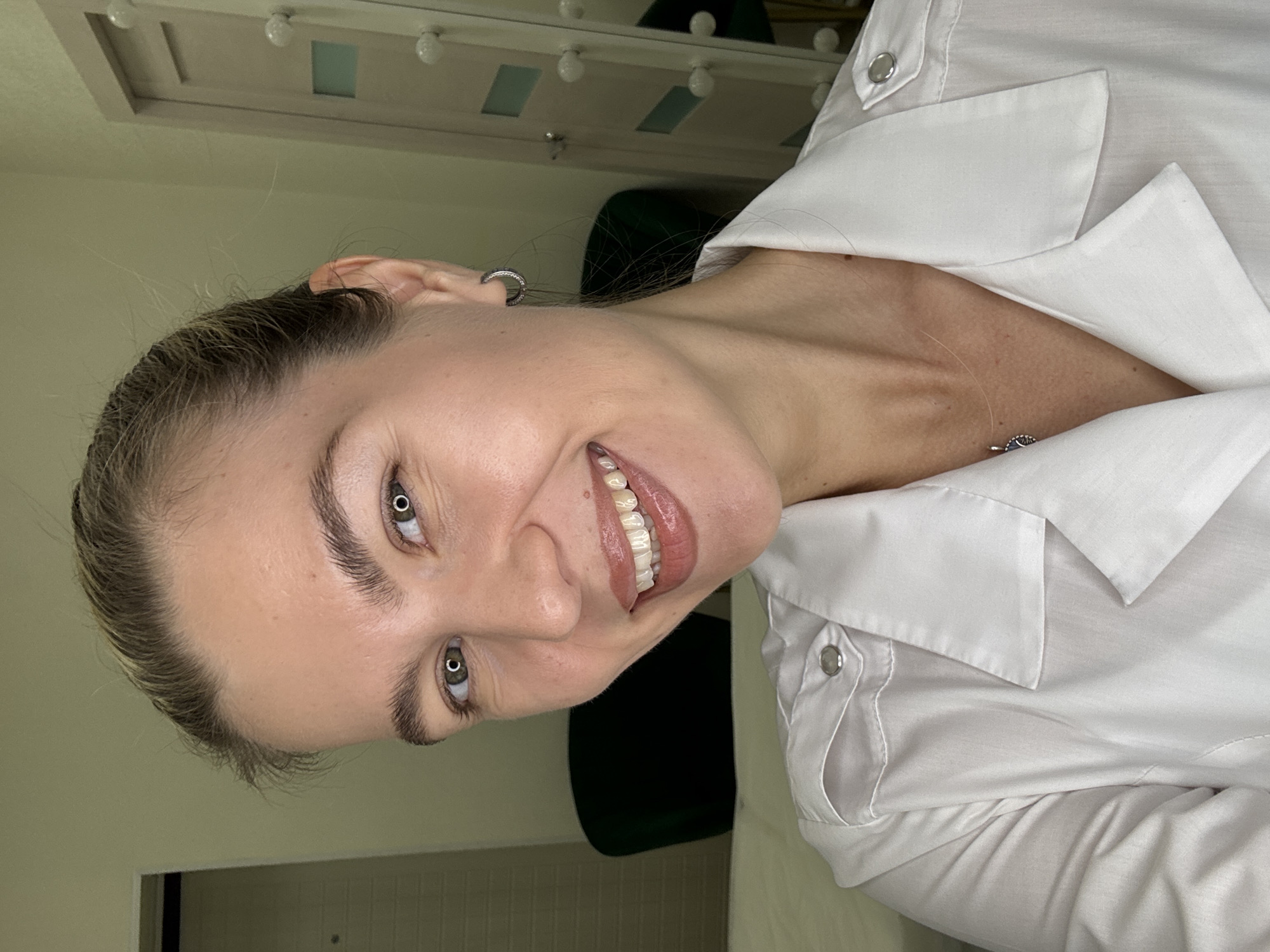 A young Caucasian woman with blonde hair pulled back and green eyes is captured in a close-up, self-taken photograph, smiling directly at the camera. She is wearing a white collared shirt with silver buttons on the shoulders and a delicate silver necklace with a blue pendant. She has a small hoop earring in her left ear. The background appears to be an indoor setting, possibly a room with light-colored walls. To the left, a portion of a dark chair is visible, and to the right, a closet or door with multiple light bulbs is partially in frame. The overall lighting suggests an artificial light source.