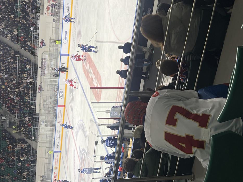 An elevated view captures an ice hockey game in progress at an indoor arena, identified as Platinum Arena in Krasnoyarsk, Russia, due to the visible "СОКОЛ" (Sokol) team logo. The ice rink occupies the central area, surrounded by spectator stands filled with numerous attendees.
On the ice, multiple hockey players are actively engaged in play. One team wears blue and white uniforms, while the opposing team wears red and white uniforms. Players are seen skating, some holding hockey sticks, with several positioned near the boards and others in open play. Two referees, identifiable by their black and white striped jerseys, are also on the ice. Advertising banners line the perimeter of the rink, displaying various logos including "ФЕДЕРАЦИЯ ХОККЕЯ РОССИИ" (Russian Ice Hockey Federation), "ATAT", "ОлимпБет", and "БУРЭНЕРГО". A large scoreboard or display is visible in the background, showing numeric information.
In the immediate foreground, from the perspective of an upper spectator section, the back of a person wearing a white hockey jersey with large red numbers "47" and gold trim is prominent. This individual also wears a red balaclava featuring a black web pattern, consistent with the Marvel character Spider-Man. They are seated in a green stadium seat. To their left and right, other spectators are visible, some partially obscured. A metal railing separates the foreground seating area from the lower sections and the ice rink. The overall scene depicts a live sporting event with significant spectator presence.
