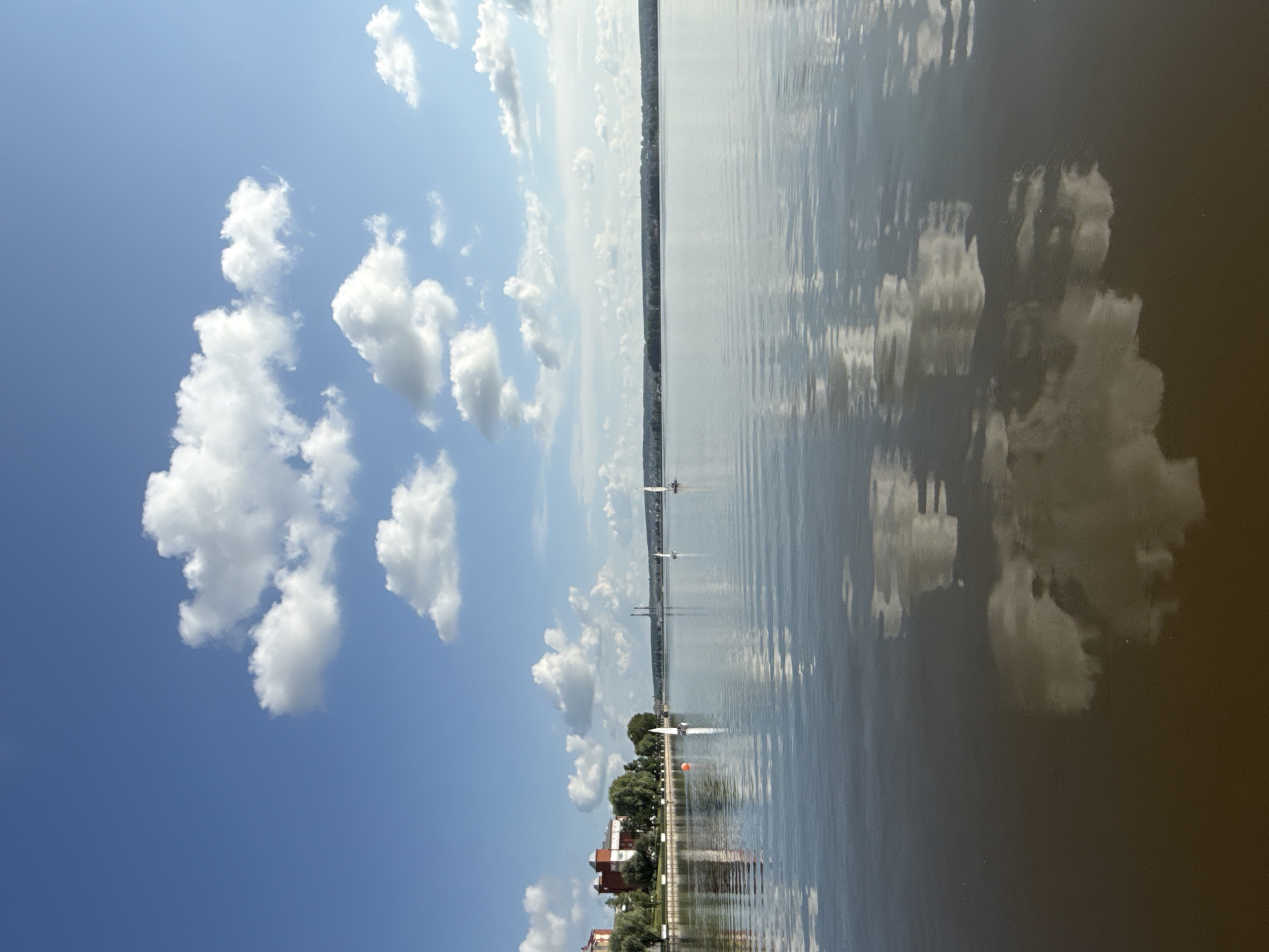 The image captures a serene panoramic view of the Votkinsk Reservoir in Votkinsk, Russia. In the foreground, the calm, dark water reflects the bright blue sky dotted with fluffy white clouds and their mirrored images. A few sailboats with white sails are visible on the water, adding a sense of gentle activity to the scene. Along the far bank, a strip of green trees lines the horizon, behind which are hints of buildings, including a prominent red-brick structure on the left. In the distance, power plant chimneys rise against the sky. The overall impression is one of tranquility and natural beauty on a clear day.