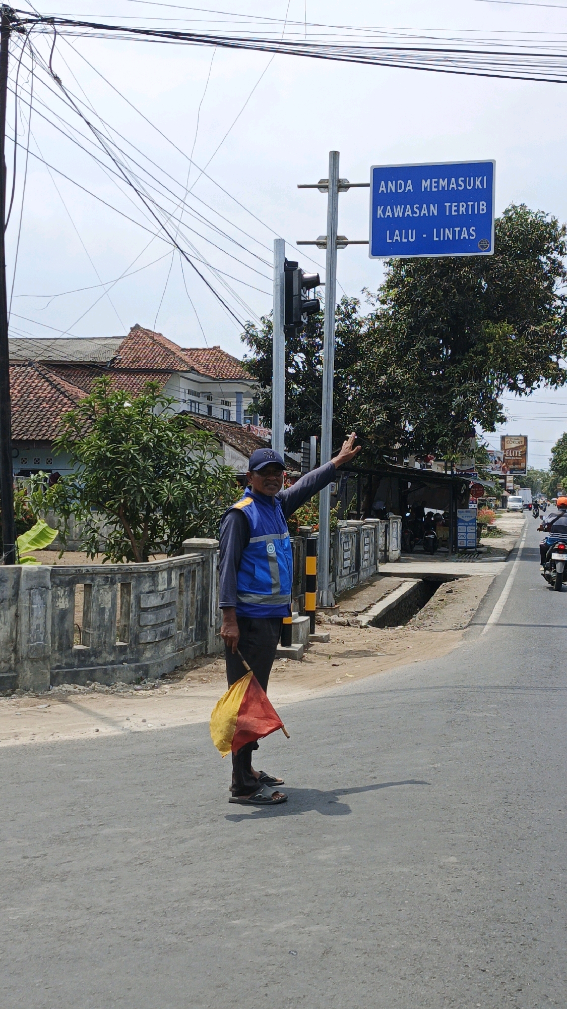 A traffic officer stands on a paved road in Desa Wonoharjo, Indonesia, directing traffic with a red and yellow flag. The officer wears a blue vest over a long-sleeved gray shirt, dark pants, a blue cap, and dark sandals. The officer's right arm is raised in a gesture of stoppage. Behind the officer, a concrete fence borders a small drainage ditch, and beyond that, trees and houses are visible. A blue sign on a pole to the right of the officer reads "ANDA MEMASUKI KAWASAN TERTIB LALU LINTAS," indicating a regulated traffic zone. Overhead power lines crisscross the sky, and in the background, motorcycles and a white van are seen on the road. The scene is under a bright, overcast sky.
