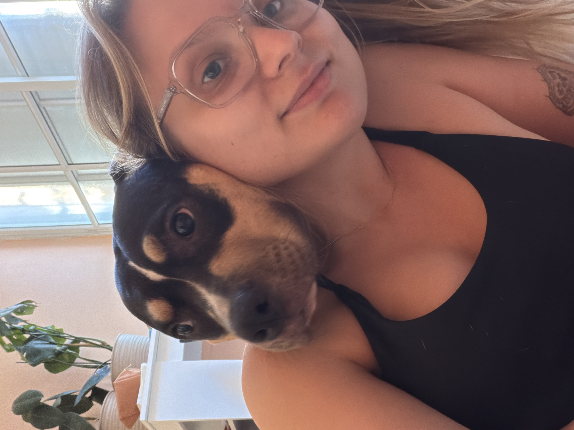 An intimate, close-up shot captures a young woman and a tri-color dog, likely indoors in Tel-Aviv, Israel, during the daytime. The woman, with light-colored hair, wears clear-framed glasses, on which the brand "CHANEL" is faintly visible on the side arm. She smiles subtly towards the camera, her face softly illuminated by bright natural light. She is wearing a black top, and a portion of a tattoo with a circular, possibly mandala-like design, is visible on her right upper arm.
Her dog, a tri-color breed with prominent black, tan, and white markings, rests its head affectionately on her left shoulder, also looking towards the camera. Its dark eyes and nose are clearly visible. In the blurred background, a window or skylight structure lets in light, and a green plant adds a touch of nature to the scene. The image conveys a feeling of warmth, companionship, and a calm moment shared between the woman and her pet.