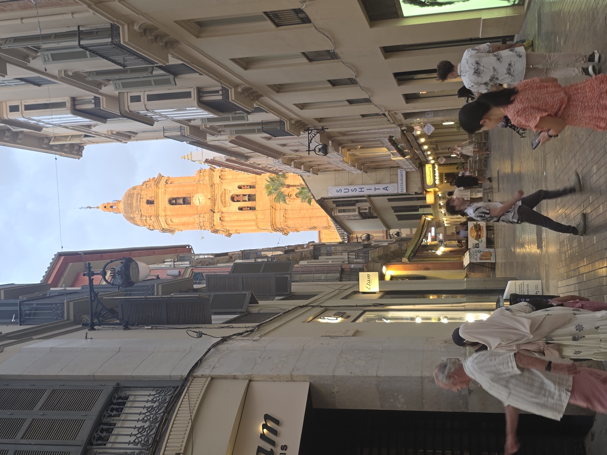 The image depicts a narrow street in Malaga, Spain, with the iconic Malaga Cathedral tower visible in the background. The tower, bathed in the warm light of what appears to be late afternoon or early evening, features a prominent clock face and bell openings. The street itself is lined with buildings exhibiting classic Mediterranean architecture, characterized by pastel-colored facades, shuttered windows, and balconies with ornate railings. Several shops and restaurants are open for business, with illuminated signs such as "Etam," "Sushita," and "Comparte" visible. People are strolling along the cobblestone street, some browsing shop windows, others seated at outdoor cafes. In the foreground, an elderly man in a light-colored shirt and pink trousers is walking towards the left, gesturing with his right hand. A woman wearing a light-colored hijab and a patterned dress, along with a child, is walking in the same direction. Further down the street, a young woman in a coral-colored dress is holding a mobile phone, and a young man in a white patterned shirt is walking away from the viewer. The overall atmosphere suggests a lively urban environment, characteristic of a Spanish city center.