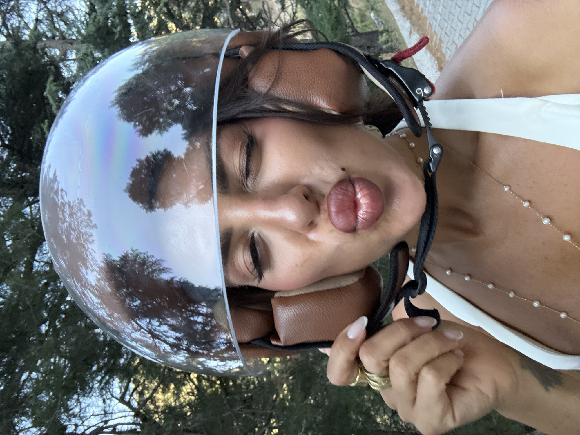 A woman wearing a motorcycle helmet with a reflective visor makes a kissy face. Her eyes are closed and her lips are pursed. She is wearing a white top and several rings on her fingers. The helmet is brown and black. The background is blurred trees. This photo was taken in Fethiye, Turkey.