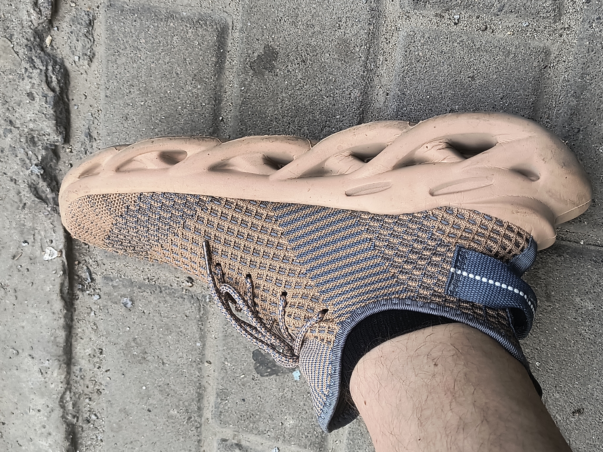 A close-up, eye-level shot shows a person's right foot wearing a tan and blue knit athletic shoe with a thick, contoured, tan sole. The shoe is on a gray, textured concrete sidewalk with visible cracks and debris. The person's bare leg, covered in light hair, is visible above the ankle. The shoe is positioned to the right of the frame, with the laces untied and hanging loosely. The lighting suggests it is daytime. The scene is devoid of any identifying markers of Rivne, Ukraine.