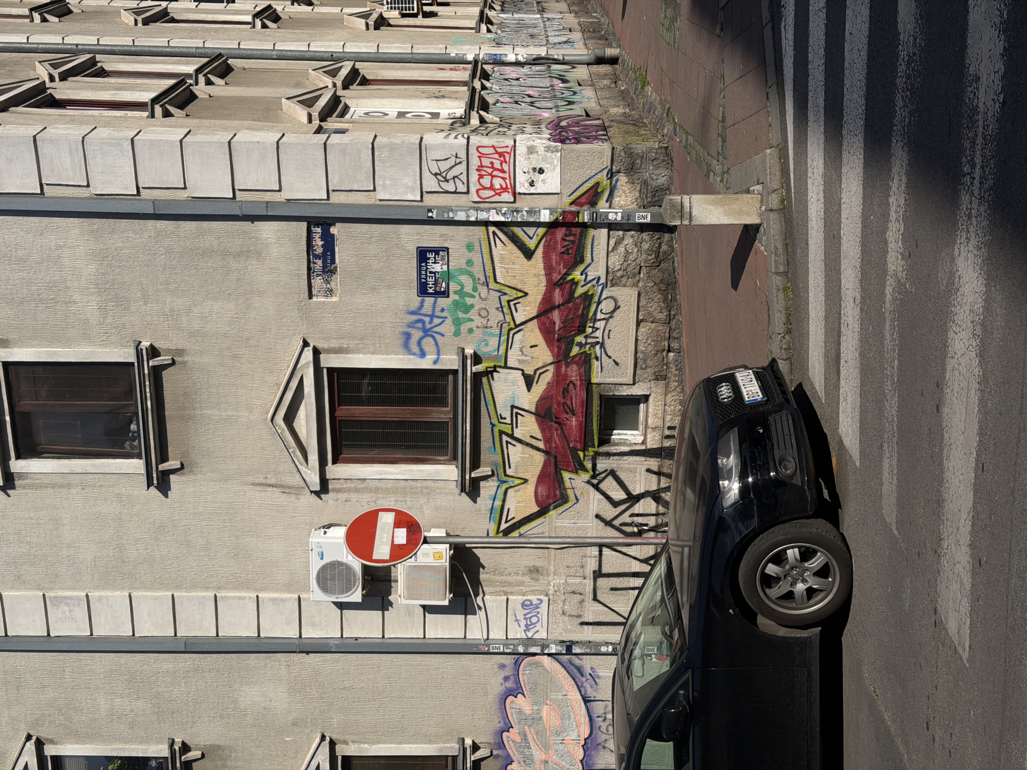 A street scene in Belgrade, Serbia, shows a building façade with graffiti, two visible windows, and a "no entry" sign attached to an air conditioning unit. A black car is parked next to a pedestrian crossing. The street is paved, and the building is beige with textured walls. Street signs in Serbian are visible.