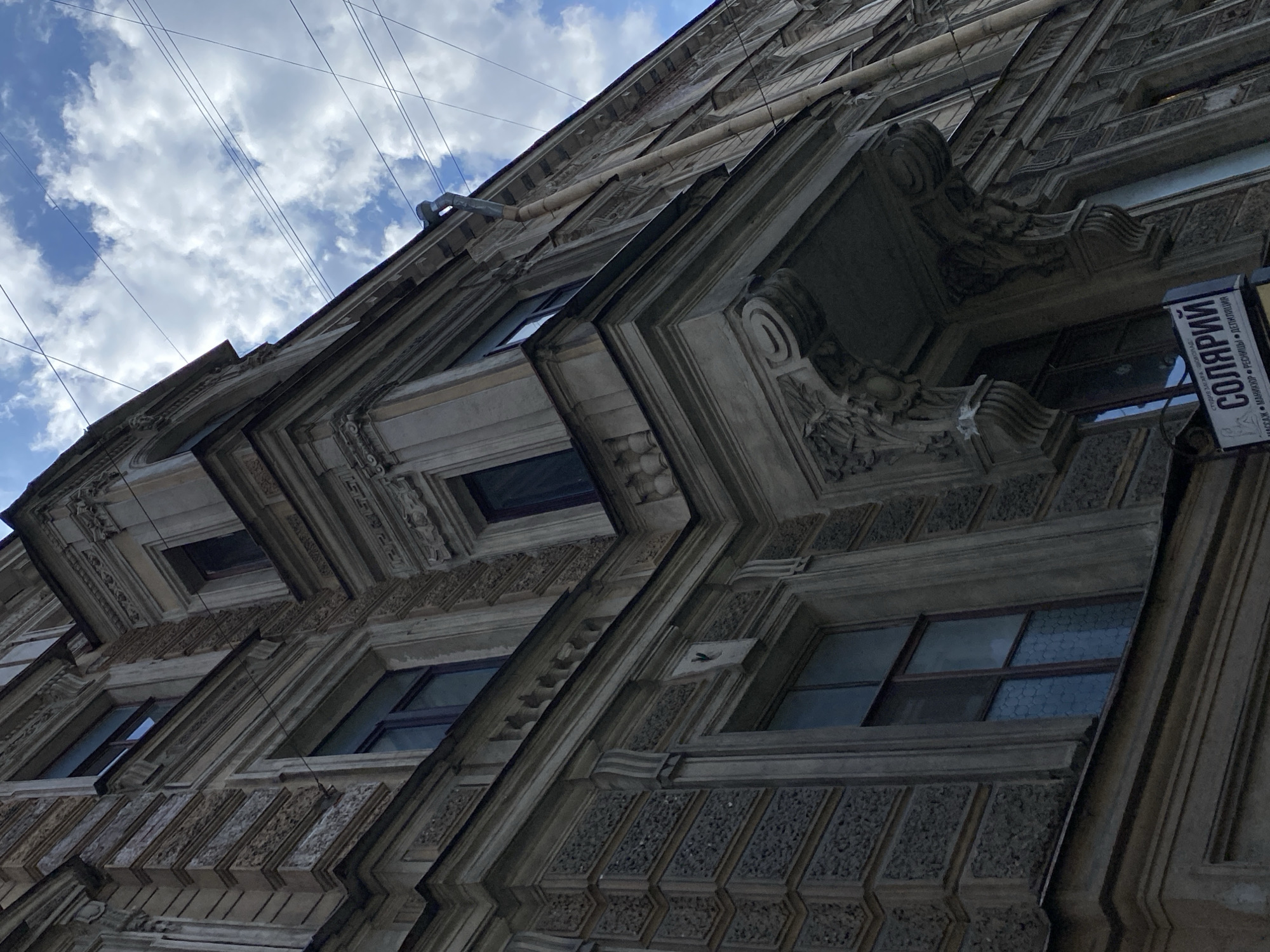 This is a low-angle, close-up shot of the ornate facade of a historical building in Saint Petersburg, Russia. The building features intricate architectural details such as cornices, decorative moldings, and textured stonework around the windows. Several windows, some with reflective glass, are visible. A balcony with elaborate corbels and a curved overhang is a prominent feature. A downspout runs vertically along the side of the building. Above the building, the sky is partly cloudy with a bright blue sky visible. Power lines crisscross the sky. A sign is partially visible on the lower left side of the image, with Cyrillic text that appears to read "СОЛЯРИЙ" and "ПАССАЖ". This sign indicates that the building likely houses a solarium or a commercial establishment. The overall impression is one of aged grandeur and urban detail within the historic center of Saint Petersburg.