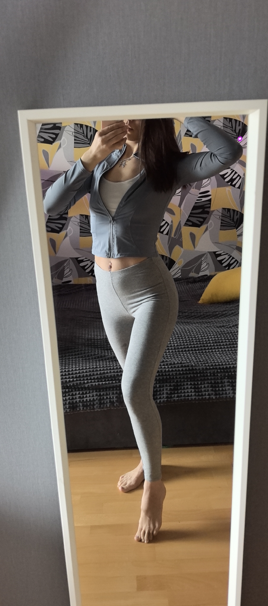 A woman stands in front of a mirror, taking a photo of herself with her phone. She is wearing a light blue zip-up jacket, a white top underneath, and grey leggings. Her hair is dark, and she is wearing a necklace and a navel piercing. The background features a bed with a patterned headboard and a grey blanket. The floor is wooden, and the walls are painted in a neutral tone. The location is Unknown, Unknown.