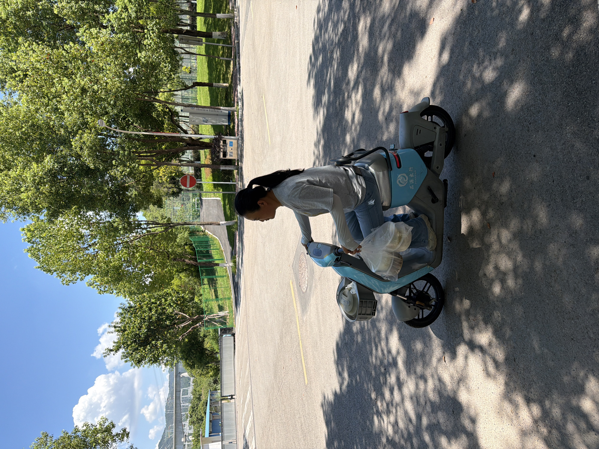 A woman with dark hair pulled back in a ponytail, wearing a grey t-shirt, blue jeans, and grey shoes, rides a blue electric scooter on a paved surface. She is carrying a clear plastic bag filled with white containers in her right hand. The scooter has a basket at the front and a logo with Chinese characters on its rear panel. The scooter is casting a shadow on the ground, which is also dappled with shadows from overhead trees. In the background, there are more trees, a green fence, a red and white circular sign, and a light pole. A bridge and some buildings are visible in the distance under a clear blue sky with fluffy white clouds. The overall setting appears to be an outdoor area, possibly a park or a campus.