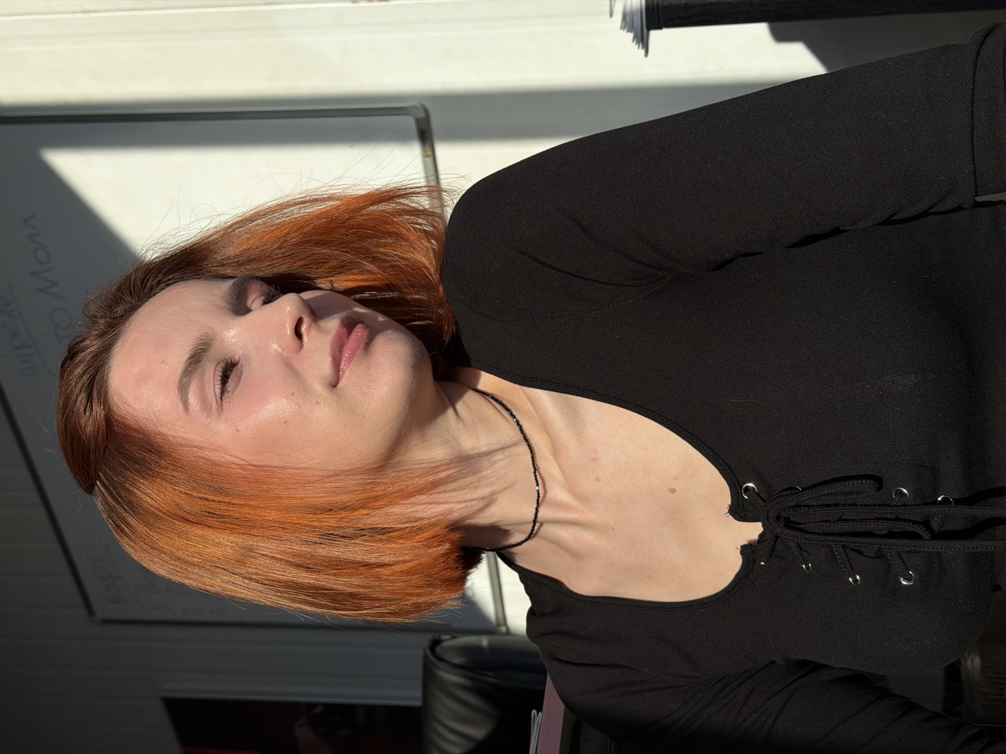 A woman with light skin and reddish-orange hair is reclining, possibly outdoors given the bright lighting. She is wearing a black long-sleeved top with a lace-up detail at the neckline and a delicate black necklace. Her eyes are closed, and her expression is serene. In the background is a white surface with some writing on it, and a glimpse of a black chair. There is no information in the image to identify the location as Chaikovsky, Russia.