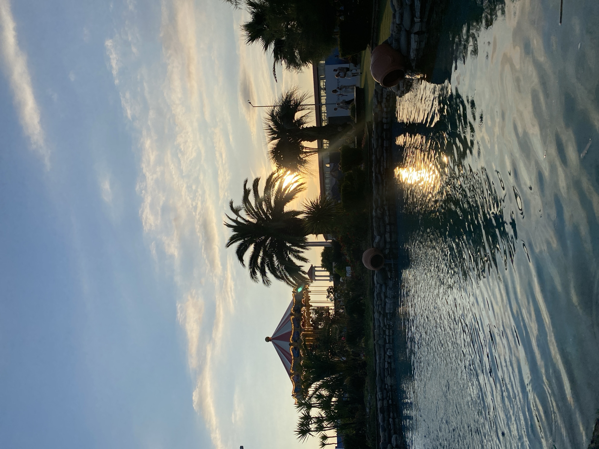 The image depicts a serene outdoor scene with a body of water in the foreground, reflecting the golden hues of the setting sun. Palm trees frame the view, their silhouettes stark against a sky adorned with wispy clouds. In the background, a carousel, illuminated by warm lights, hints at a festive atmosphere. A building with visible text, possibly a hotel or resort, stands behind the carousel, suggesting this is a leisure or entertainment area. The time of day is clearly sunset, with the low sun casting long shadows and creating a tranquil, golden-hour glow. The presence of a building and a carousel in proximity to palm trees and water suggests a location in a warm climate, possibly a resort town. The scene evokes a sense of peace and relaxation, with the gentle ripples on the water adding to the calm ambiance. The overall impression is one of a picturesque evening in a holiday destination.