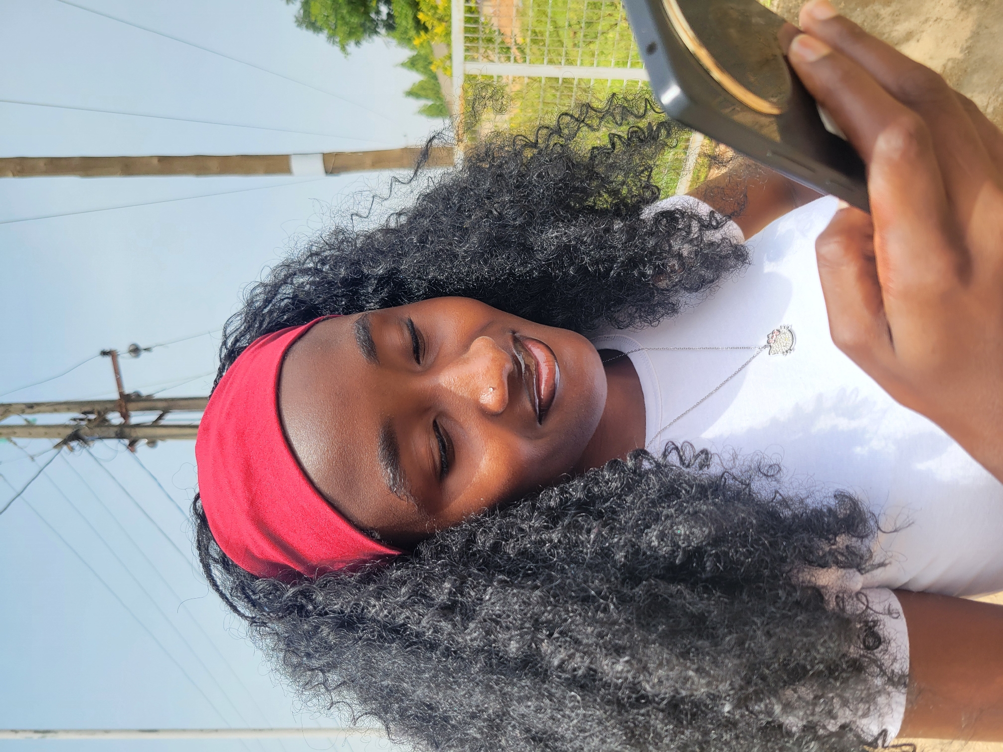 A medium shot presents a young Black woman with tightly curled, dark hair, accented by a red headband. Her eyes are closed, and she has a nose piercing. She is wearing a white t-shirt and a silver necklace with a pendant. Her right hand holds a cell phone up towards her face. The background shows a blue sky, utility poles with electrical wires, a partial view of a wire fence, and some green vegetation. The location is indicated as Jos, Nigeria.