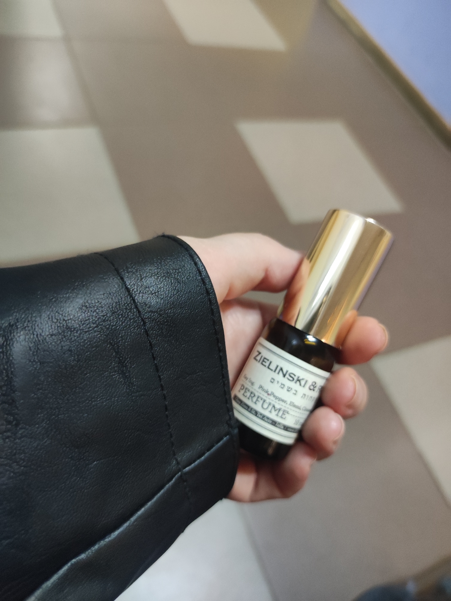 A hand clad in a black leather jacket holds a small bottle of Zielinski & Rozen perfume with a gold cap. The label on the bottle lists ingredients as "Pink Pepper, Elemi, Cloves" and states "Made and bottled in Tel Aviv - Jaffa / Israel." The background appears to be a tiled floor and a wall, giving an indication that it's indoor. The specific location within Votkinsk, Russia, cannot be determined from the given visual information.