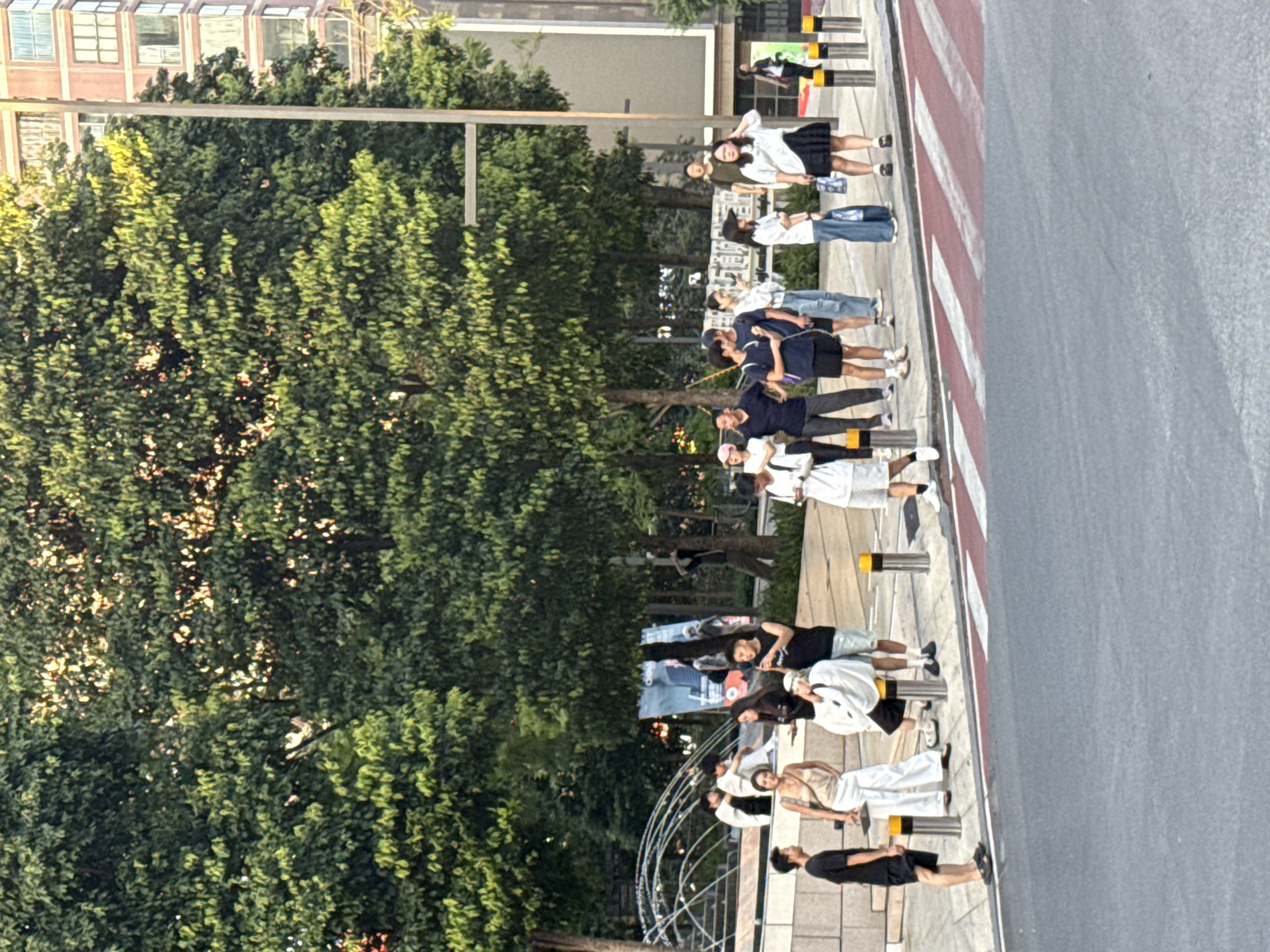 The image depicts a group of young adults standing and interacting on a sidewalk adjacent to a street in Yuzhong District, China. The scene is set against a backdrop of lush green trees and a multi-story building. Several individuals are casually dressed in t-shirts, shorts, and skirts, with some holding phones or bags. A crosswalk with red and white markings is visible on the street in the foreground. The lighting suggests it is daytime, possibly late afternoon given the sunlight filtering through the trees. A water feature with arcs of water is partially visible on the left side of the image. The overall atmosphere appears relaxed and urban.