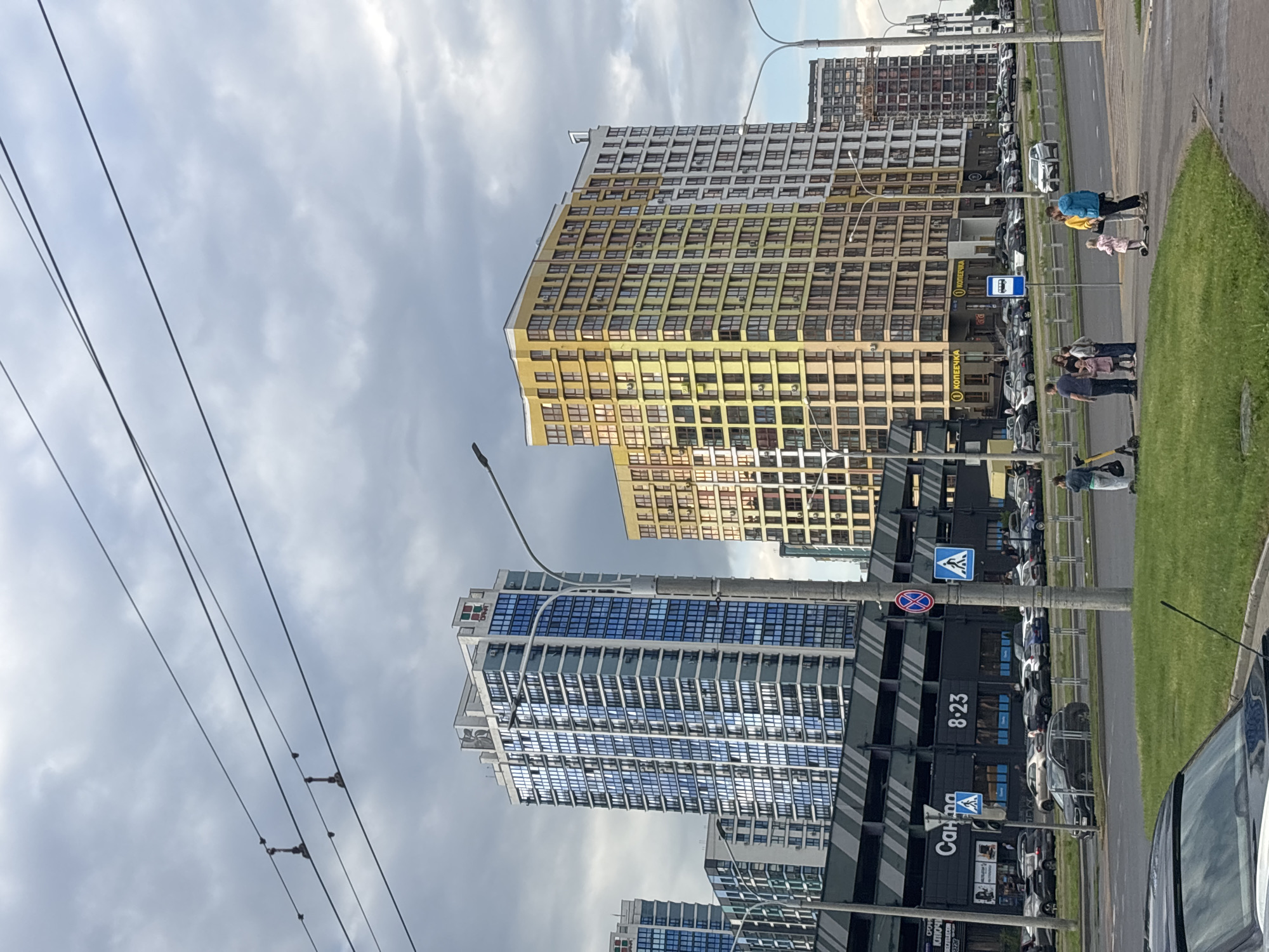 The image captures a street scene in the city of Senitsa, Belarus, featuring modern high-rise buildings and a busy road. In the foreground, a black car is partially visible on the left, with a grassy area and a sidewalk extending to the right. Several people are on the sidewalk, including a woman with a backpack, a group of adults and children, and a person on a scooter. A young child is also present. Across the road, a multi-story parking garage is visible with the signage "Салона" and "8-23". Above the parking garage, several contemporary apartment buildings dominate the skyline, with one prominent blue-windowed tower and a yellow and beige multicolored tower. Further in the distance, more apartment buildings can be seen. Overhead, power lines stretch across the cloudy sky. The environment appears to be urban, with evident infrastructure for transportation and housing.