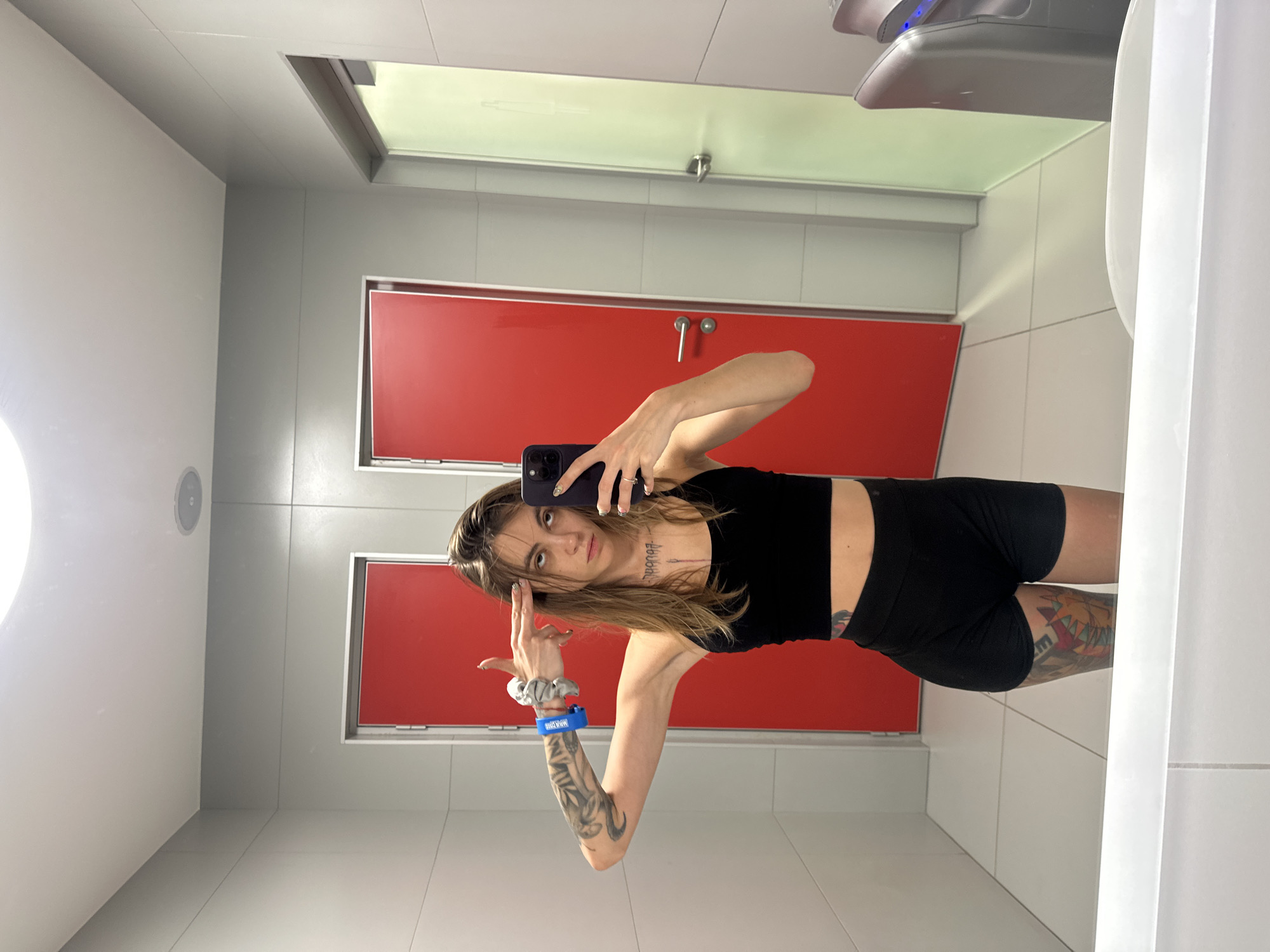 A young woman with long brown hair, wearing a black tank top and black shorts, stands in a brightly lit, modern restroom and takes a selfie in a large mirror. She has multiple tattoos on her arms and legs. Her right hand is raised to her head, with her index finger extended and touching her temple, while her thumb forms a gun shape. She is looking directly into the camera with a neutral expression. The restroom has white tiled floors and light gray walls. Two red doors are visible behind her, along with a light green door to the right. A hand dryer is also visible on the right side of the frame. The scene appears to be in a public establishment. There is no visual information within the image to indicate that this location is in Moscow, Russia.
