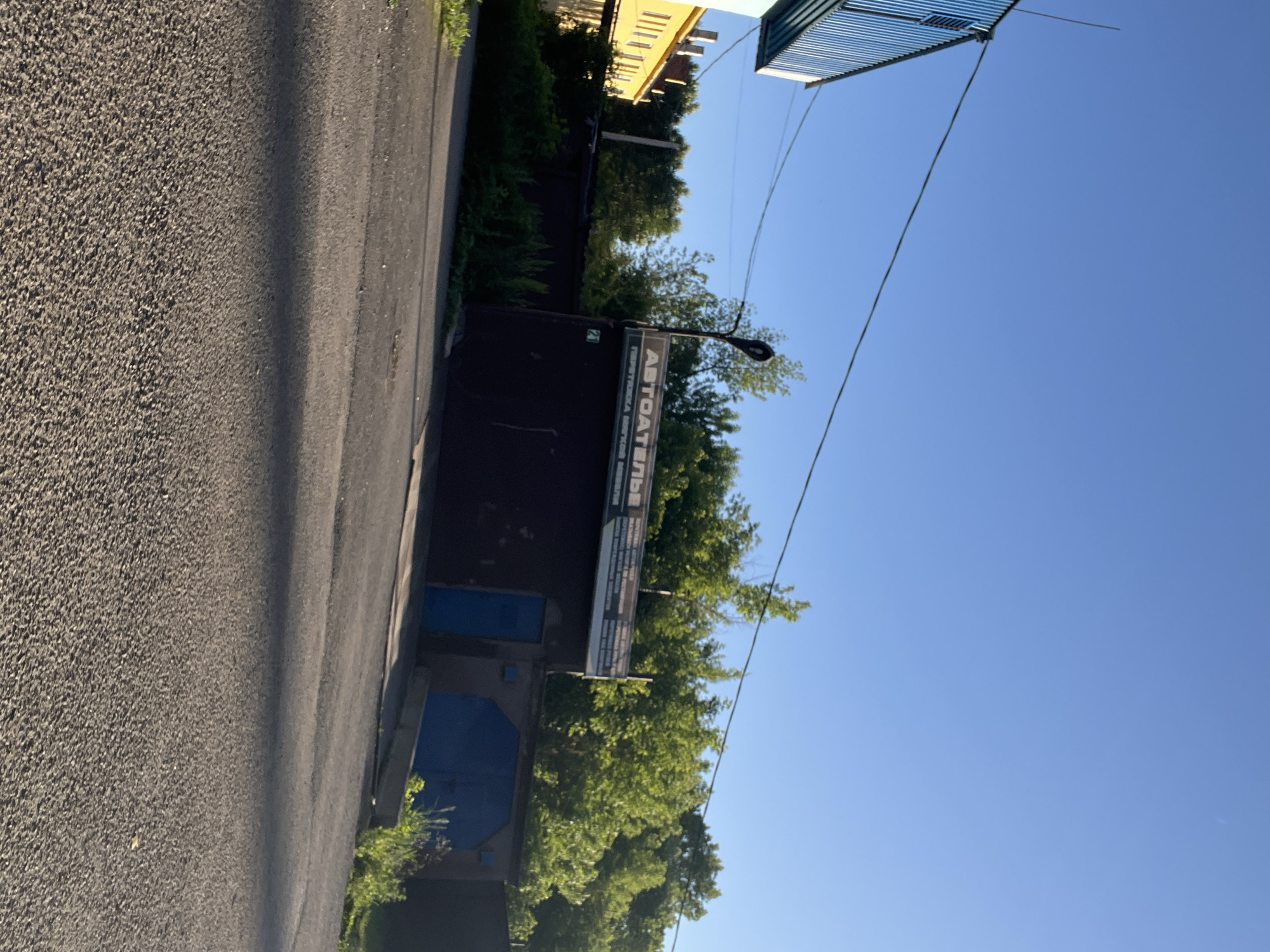 This media file depicts an outdoor scene under a clear blue sky, suggesting it was taken during daylight hours with fair weather. The composition is oriented sideways. In the foreground, a dark asphalt road with a textured surface runs along the left side of the frame.
To the right of the road, a building with a dark facade is visible. On this building, there is a sign with text that appears to be in Russian. The visible portion of the text reads "АВТОАТЕЛЬЕ" followed by more text in a smaller font. Above the building, lush green trees are abundant, creating a natural backdrop.
Further in the background, a yellow building with multiple windows and chimneys can be seen, possibly residential or administrative. A blue corrugated metal structure, possibly a roof or awning, is present in the upper right portion of the image. Overhead, electrical wires are strung across the sky, one of which runs diagonally through the frame. The overall setting appears to be an urban or semi-urban environment, likely a street or alleyway in Kiselyovsk, Russia, given the provided location context. There are no people visible in the image. The scene conveys a sense of everyday life in this location.